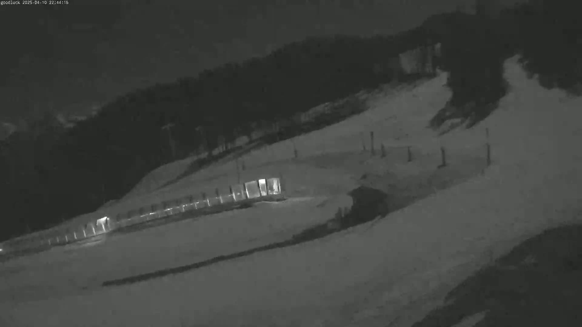 A nighttime, snow-covered ski slope with a fence and a structure illuminated in the lower left corner.