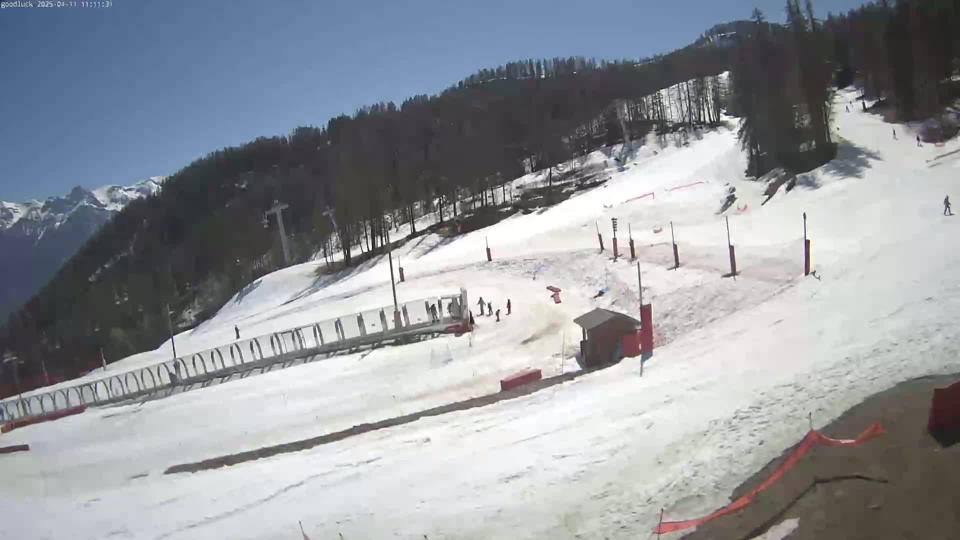 A snow-covered ski slope with several skiers and snowboarders, a ski lift, and a small building is visible under a clear blue sky.