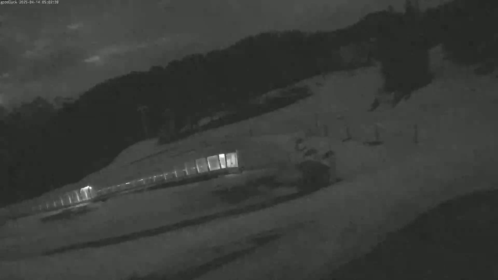 A dimly lit, nighttime scene shows a long structure with illuminated windows situated on a snow-covered slope beneath a dark sky.