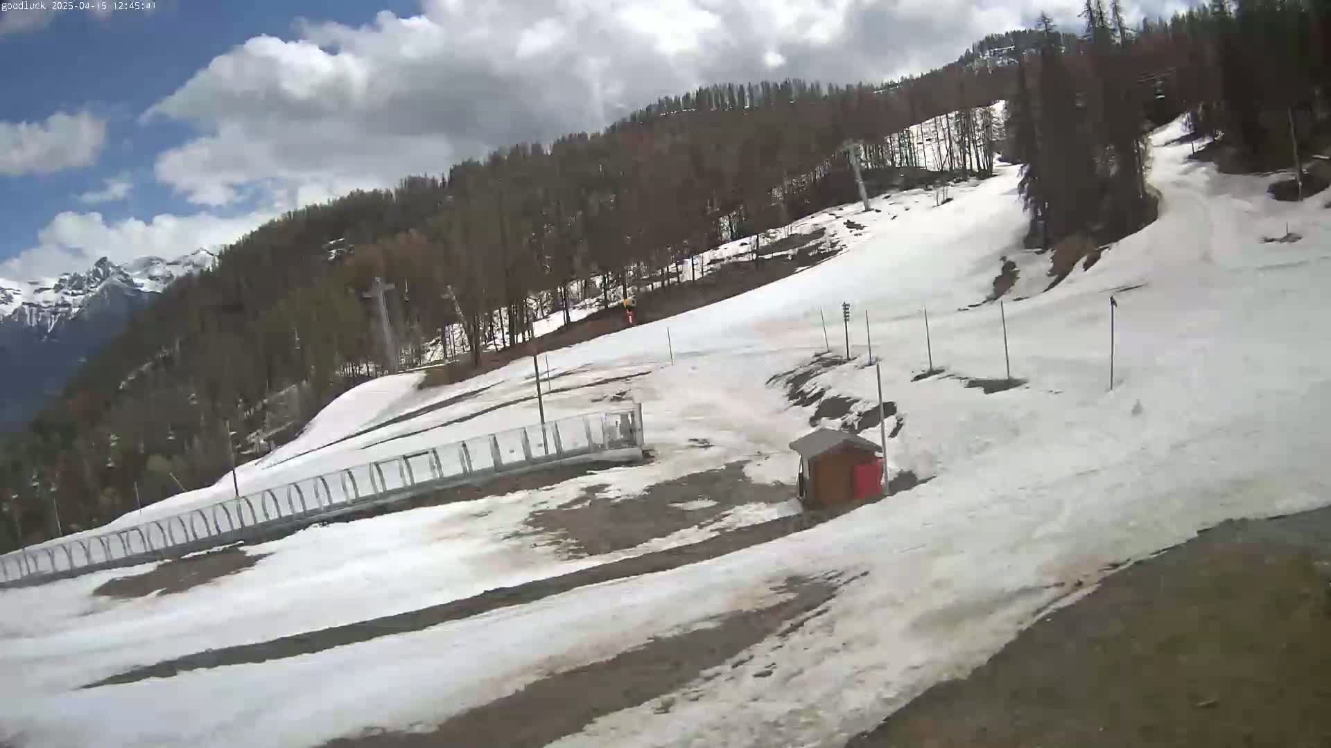 A partly cloudy day reveals a snow-covered ski slope with a small wooden structure and a metal fence, nestled among a coniferous forest and distant mountains.