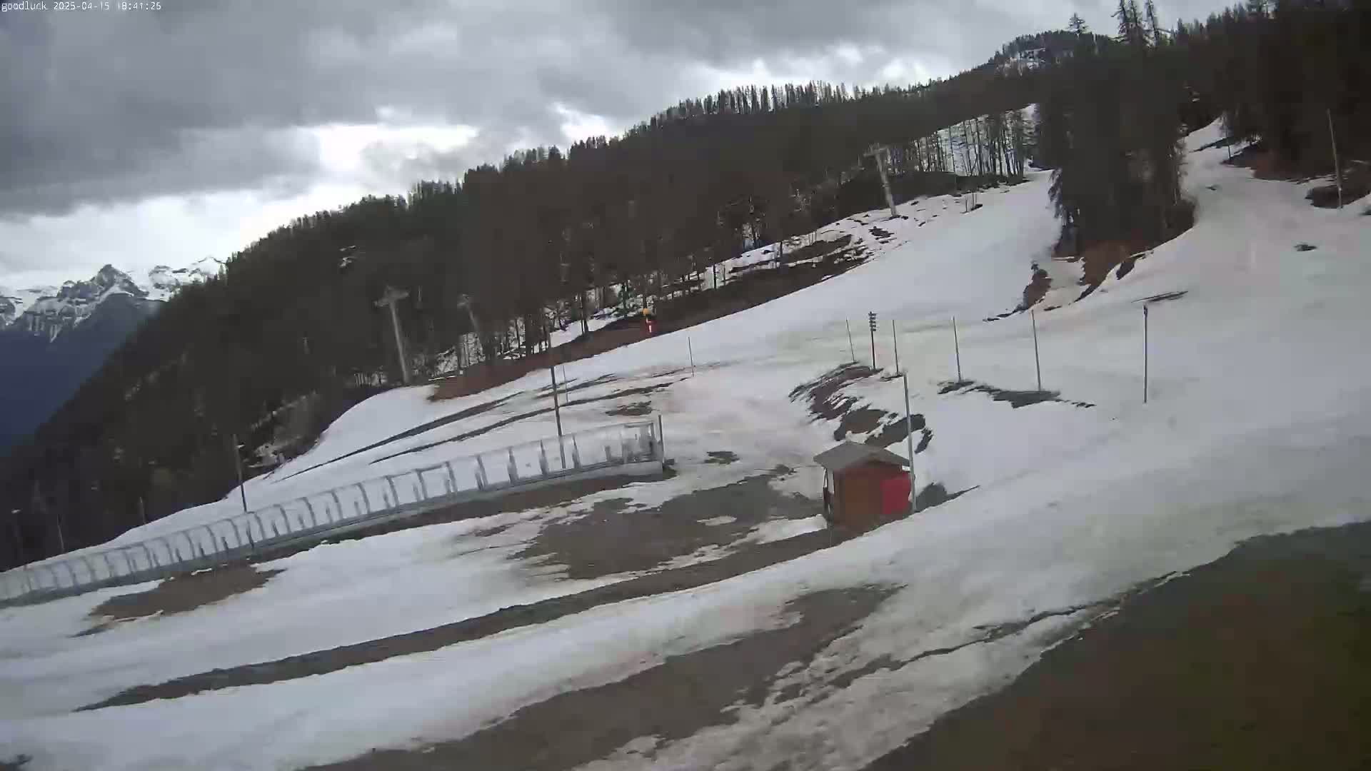A mostly cloudy day reveals a snow-covered ski slope with a small building and a metal fence, nestled amongst a dark green forested mountainside.