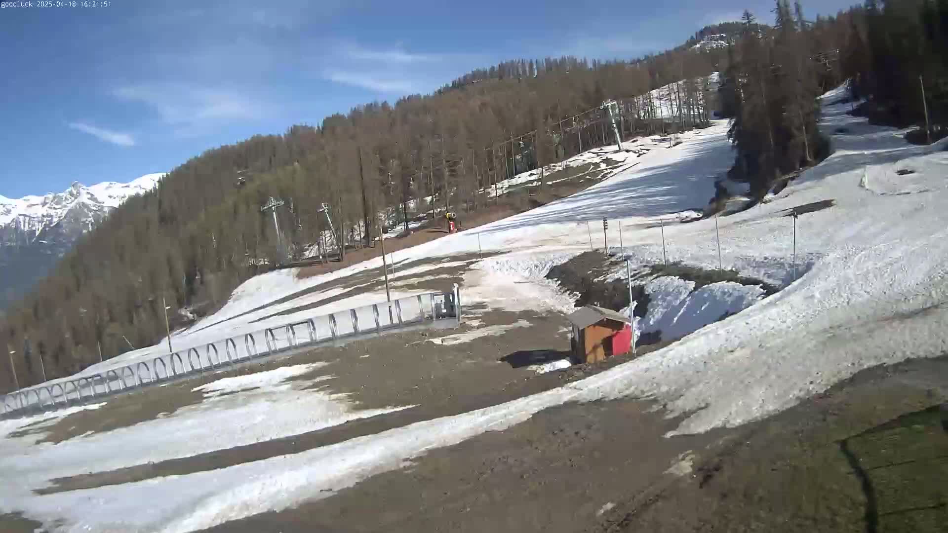 A mostly sunny day reveals a ski slope with patches of melting snow, a ski lift, and a small building, set against a backdrop of snow-capped mountains and a forest.