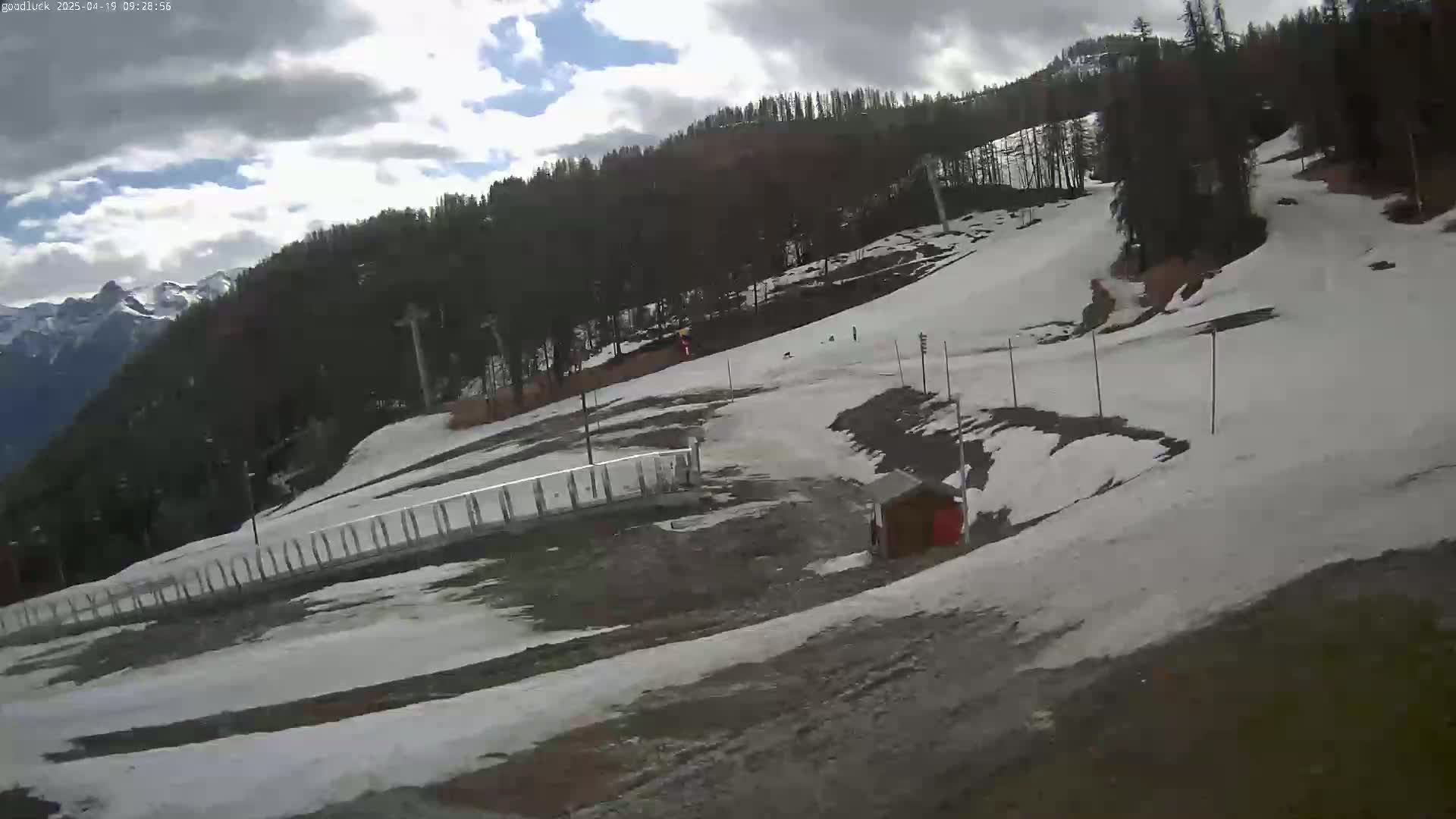 A partly cloudy day shows a snowy ski slope with a few people visible in the distance, alongside a forested mountainside and a small building.