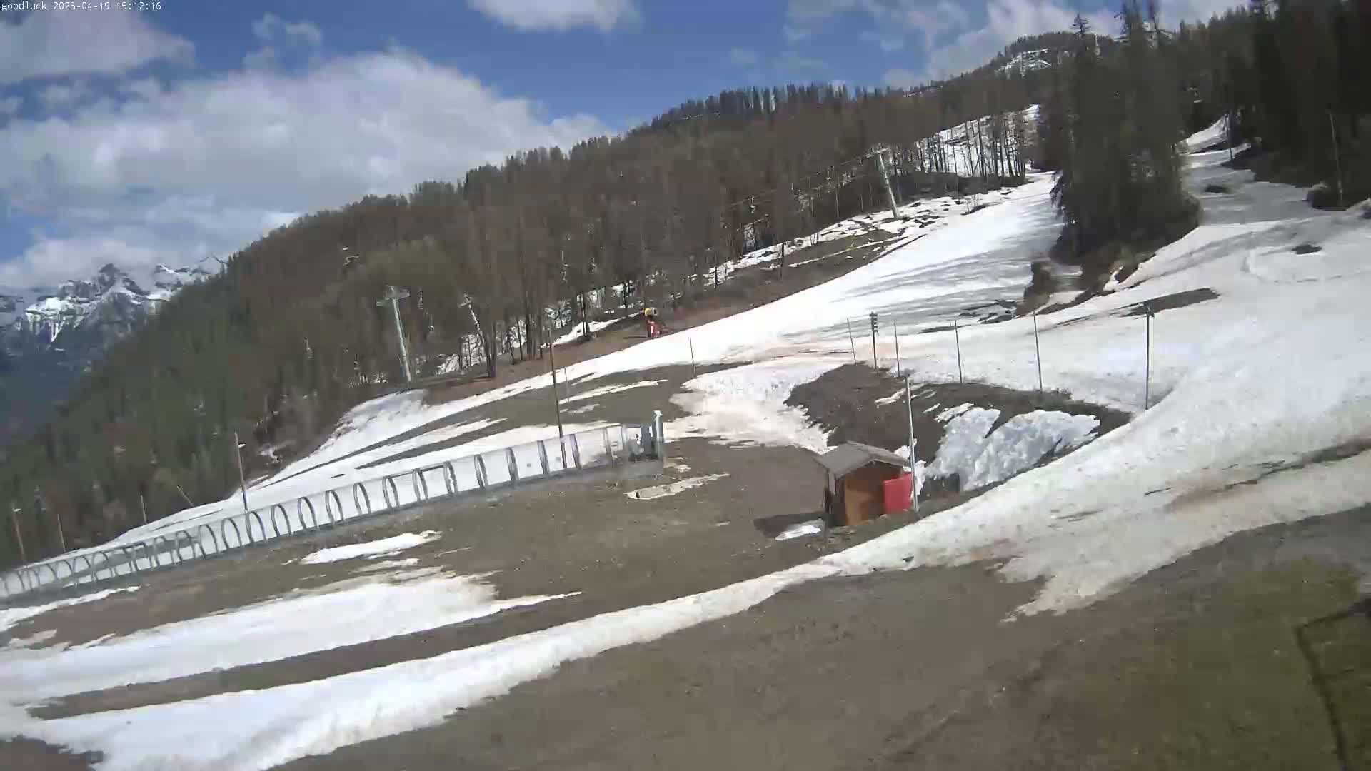 A mostly sunny day shows a ski slope with patches of melting snow, a small building, and a ski lift in a mountainous area.