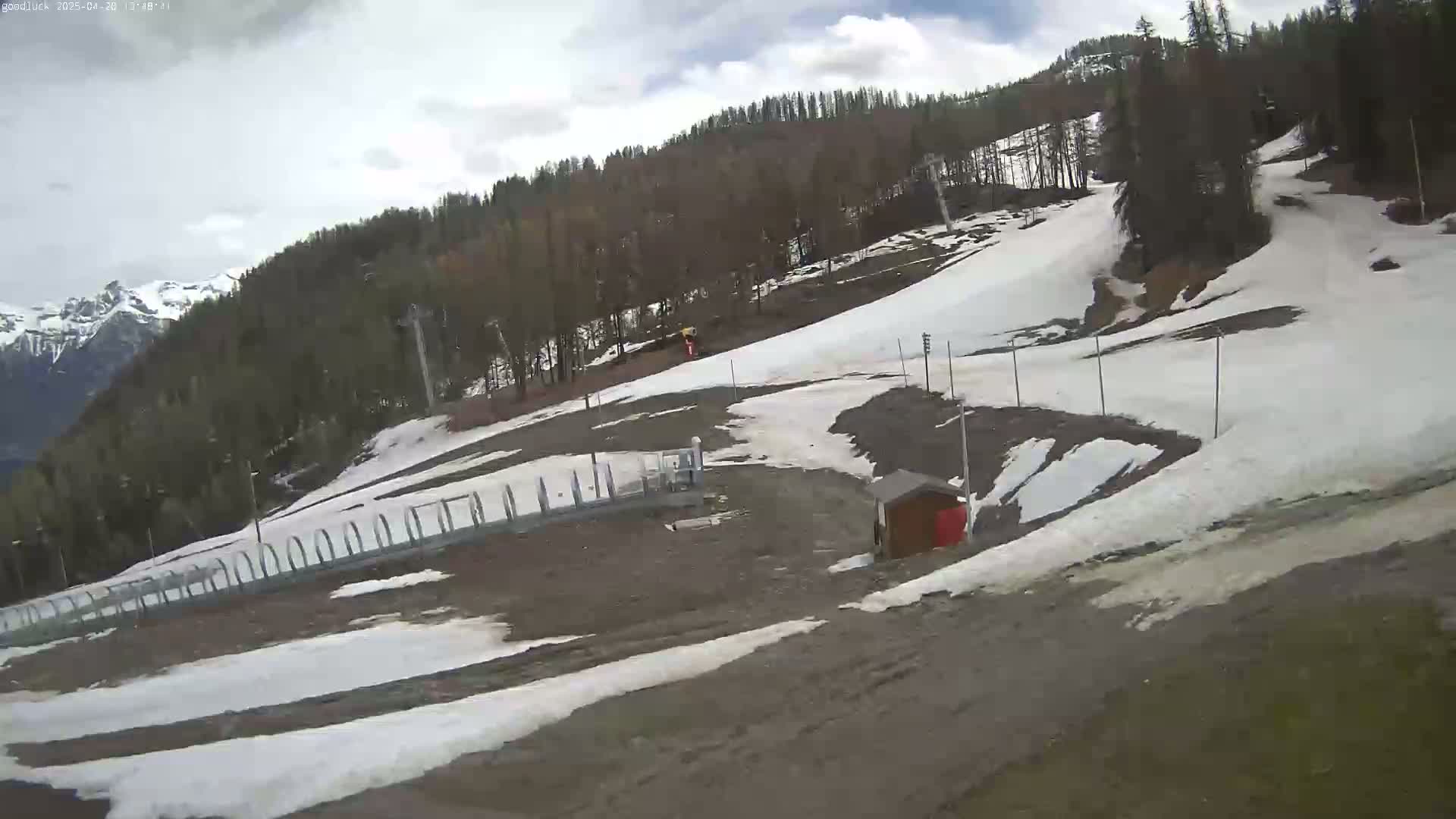 A partly cloudy day reveals a ski slope with patches of melting snow, a small building, and a fence, all set against a backdrop of a mountain range and a coniferous forest.