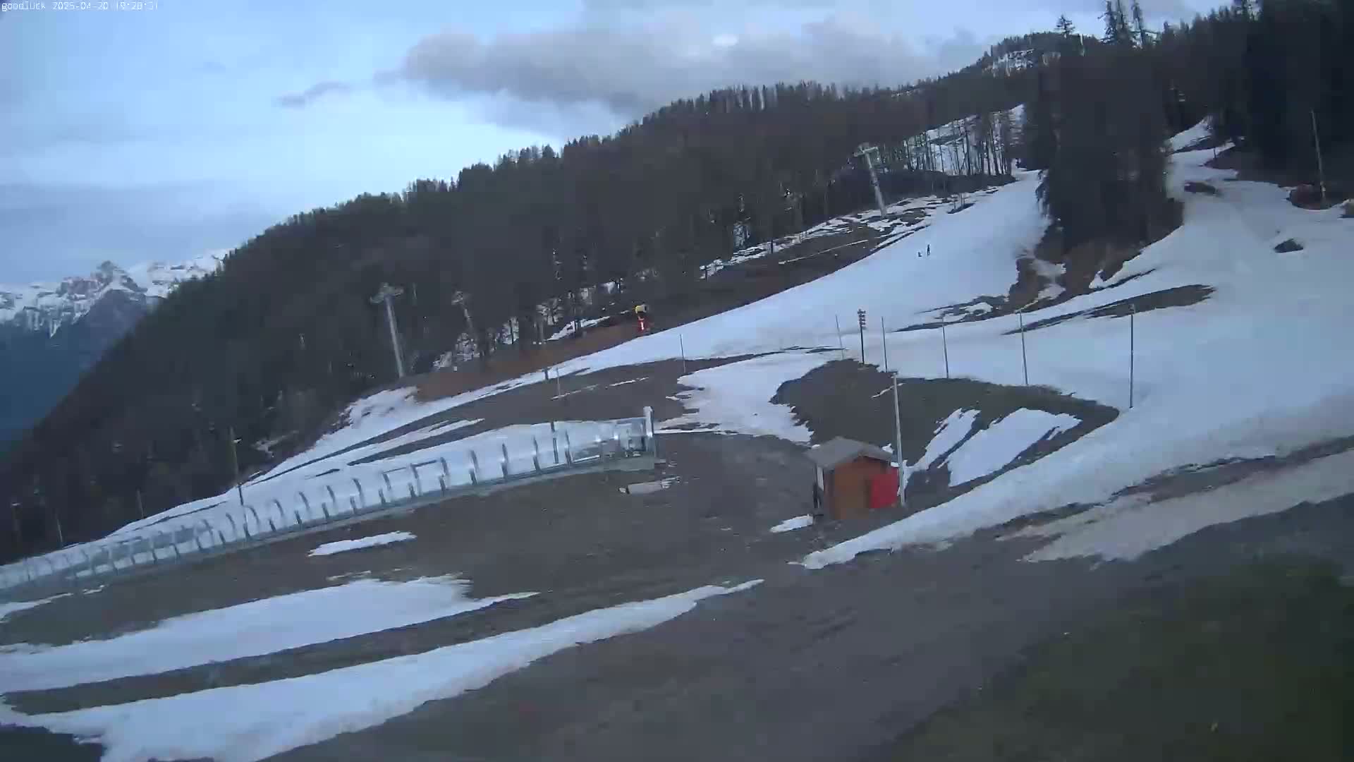 A mostly cloudy day reveals a ski slope with patchy snow, a small building, and a mountain range in the background.