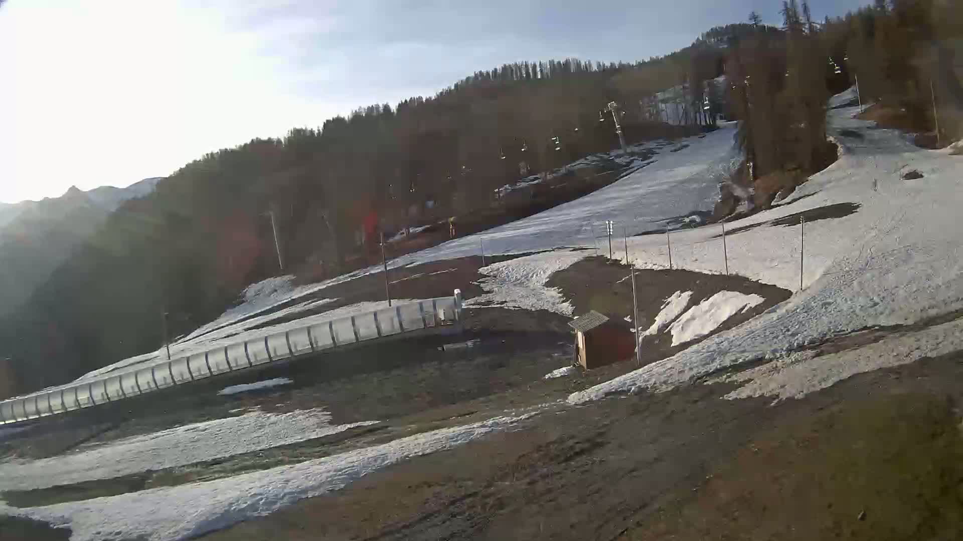 A snowy ski slope with patchy snow cover on a sunny day, featuring a long, low barrier and a small structure near the bottom.