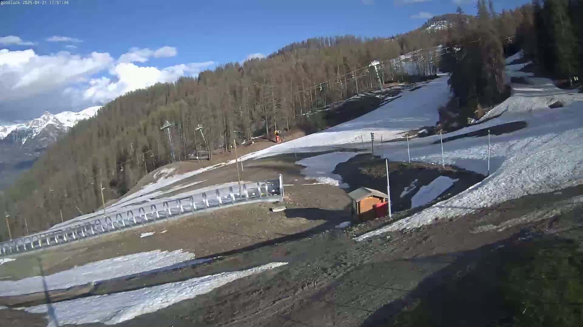 A mostly sunny day shows a ski slope with patchy snow, ski lifts, and a small building near the bottom of the hill, with mountains in the background.