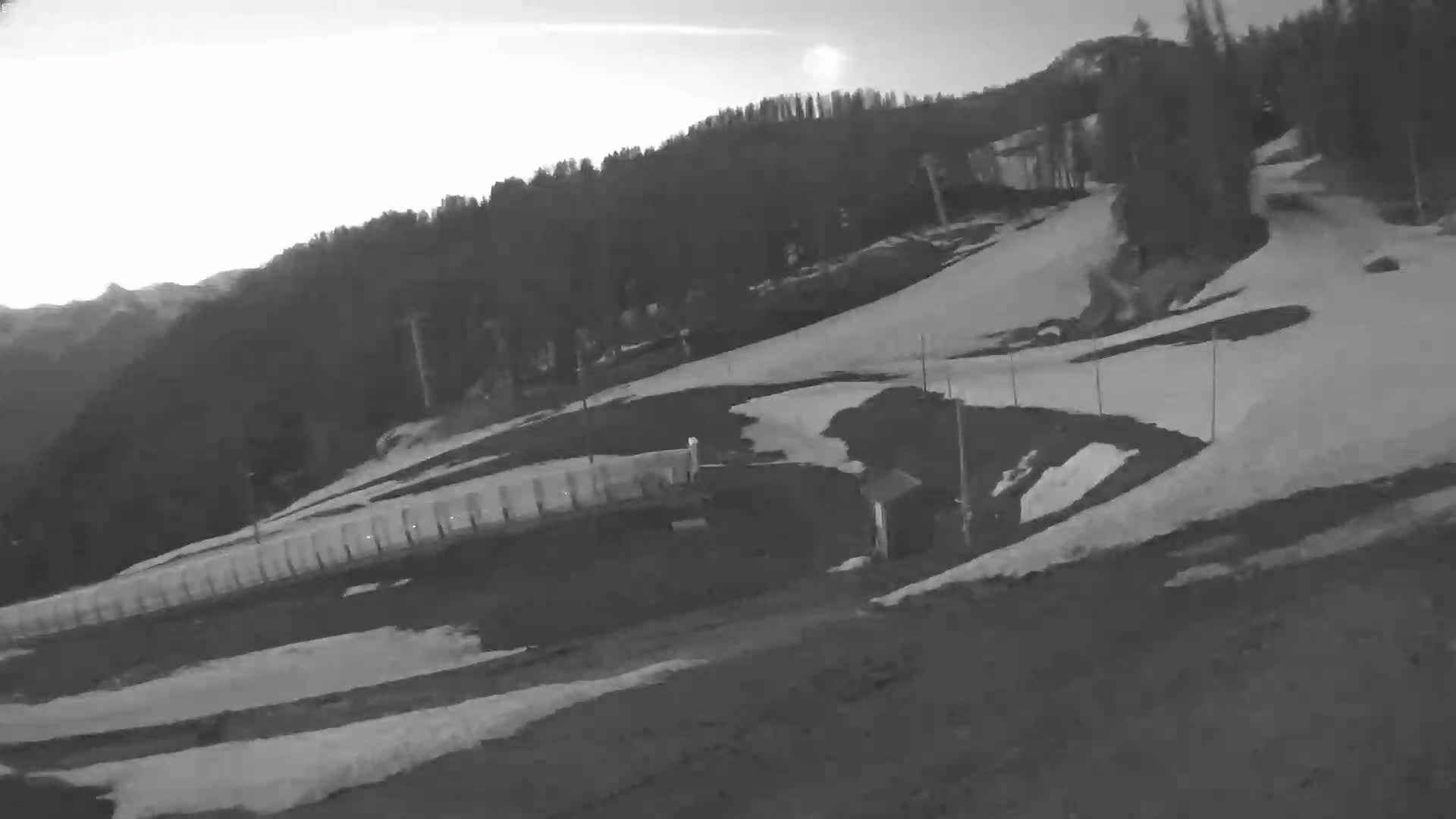 A snow-covered ski slope with a forested hillside in the background under a mostly clear sky.
