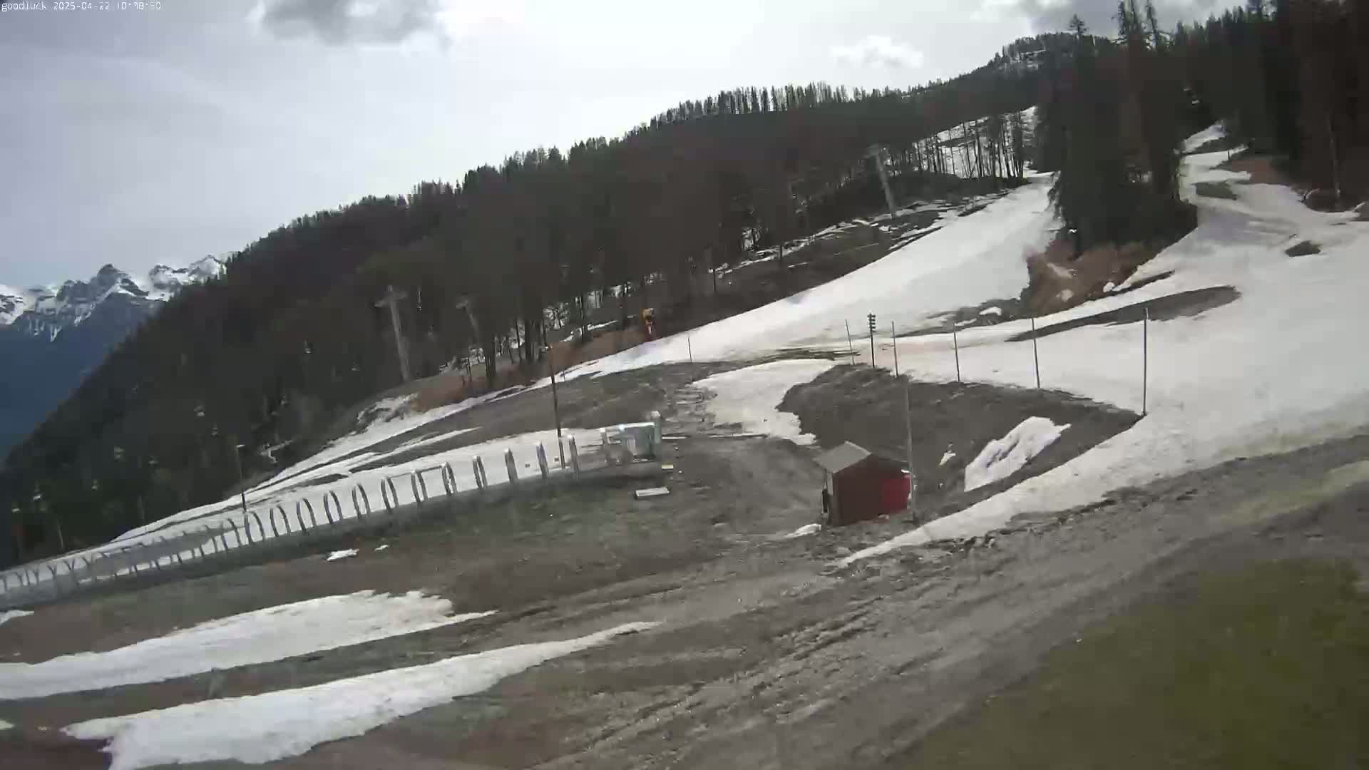 A partly cloudy day shows a ski slope with patches of melting snow, a small red shed, and a mountain range in the background.