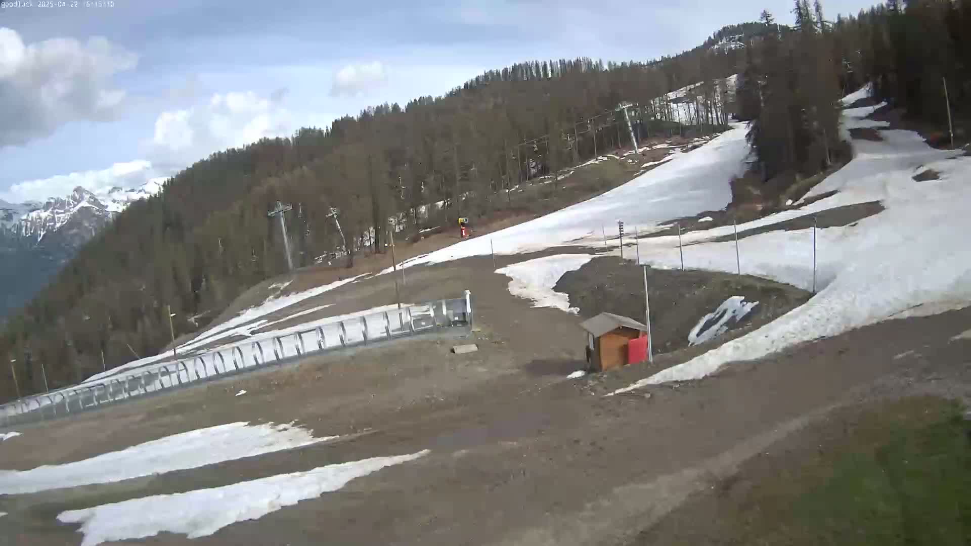 A mostly cloudy day shows a partially snow-covered ski slope with a few trees and ski lifts in the background.