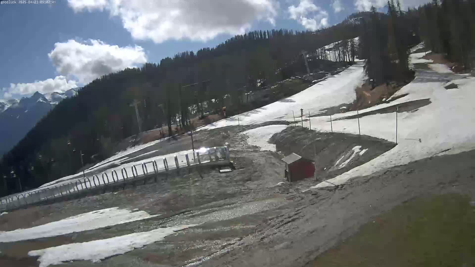 A mostly sunny day reveals a partially snow-covered ski slope in a mountainous area, with a small red building near the bottom of the slope.