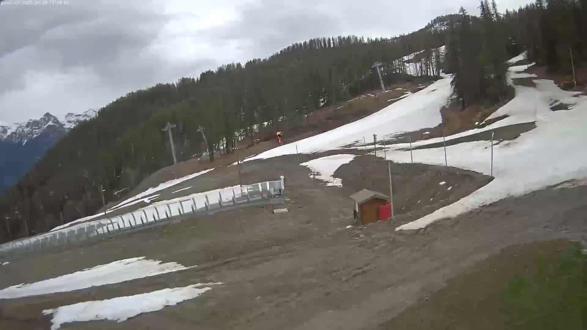 A mostly cloudy day shows a ski slope with patches of melting snow, some ski lifts, and a small building on a mountainside with a forest in the background.