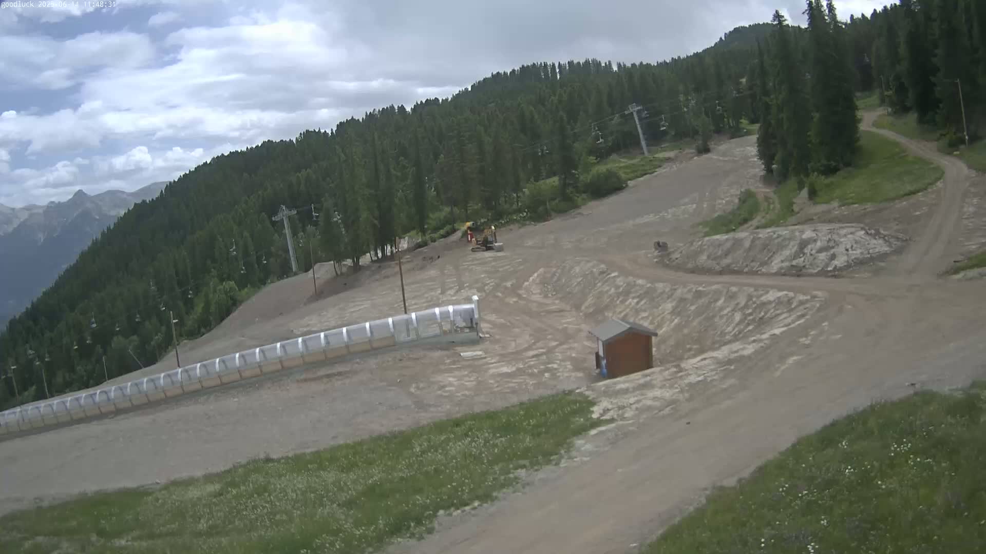 A mostly cloudy day reveals a mountain ski area under construction, showing exposed dirt, some small buildings, a long covered structure, and a small piece of heavy machinery.