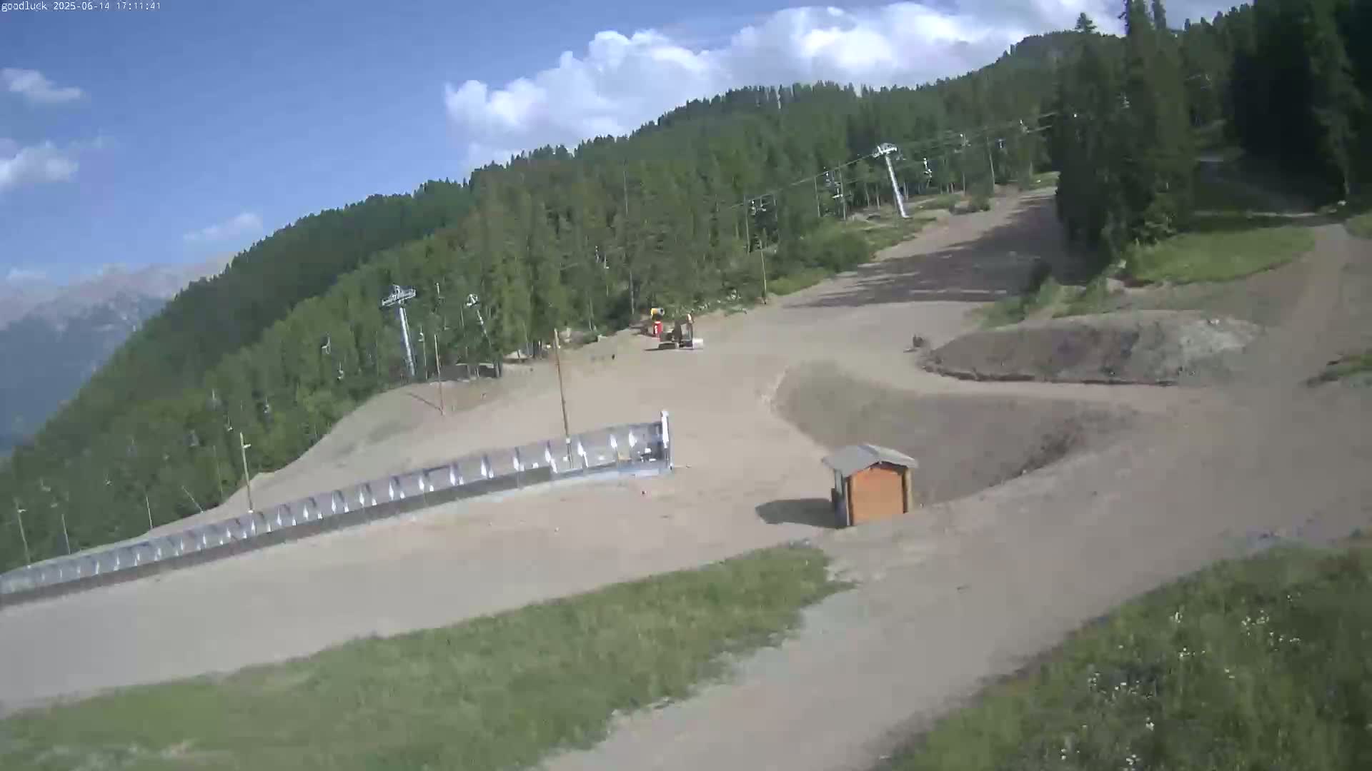 A mostly sunny day shows a mostly barren ski slope with some construction equipment, a small building, and ski lifts visible amongst the surrounding green trees and mountains.