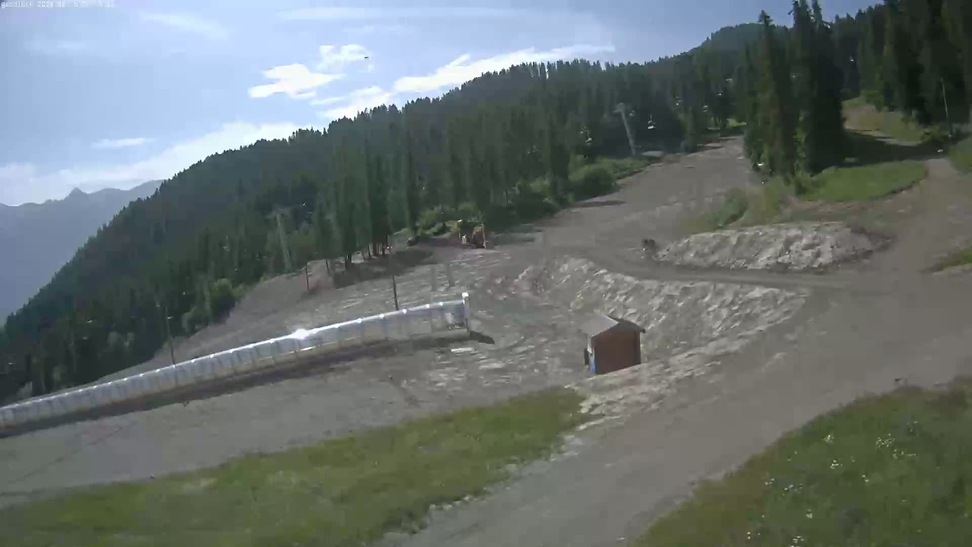 A mostly sunny day reveals a mountainside ski area with a long, covered conveyor and dirt pathways amidst evergreen trees and patchy grass.
