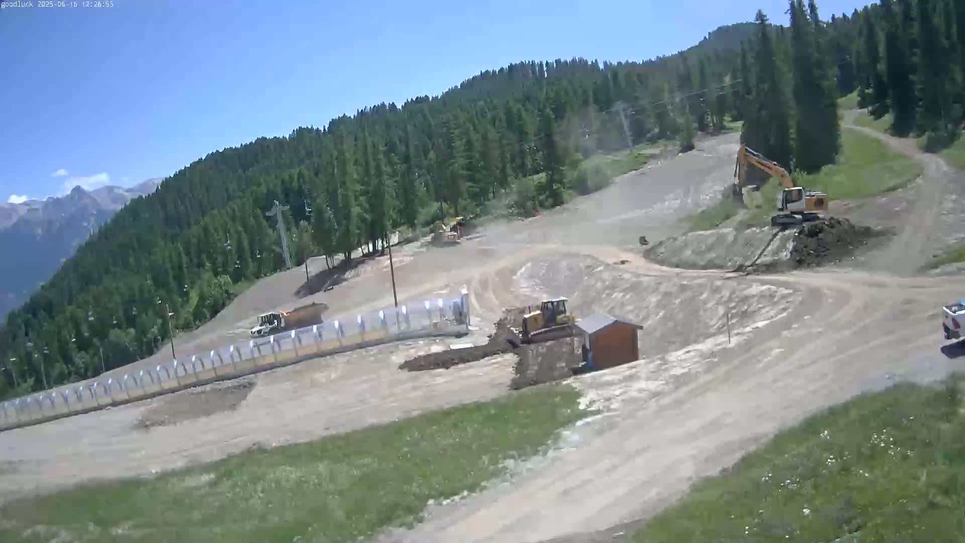 Under a clear sky, construction equipment works on a muddy hillside surrounded by a coniferous forest and mountains in the distance.
