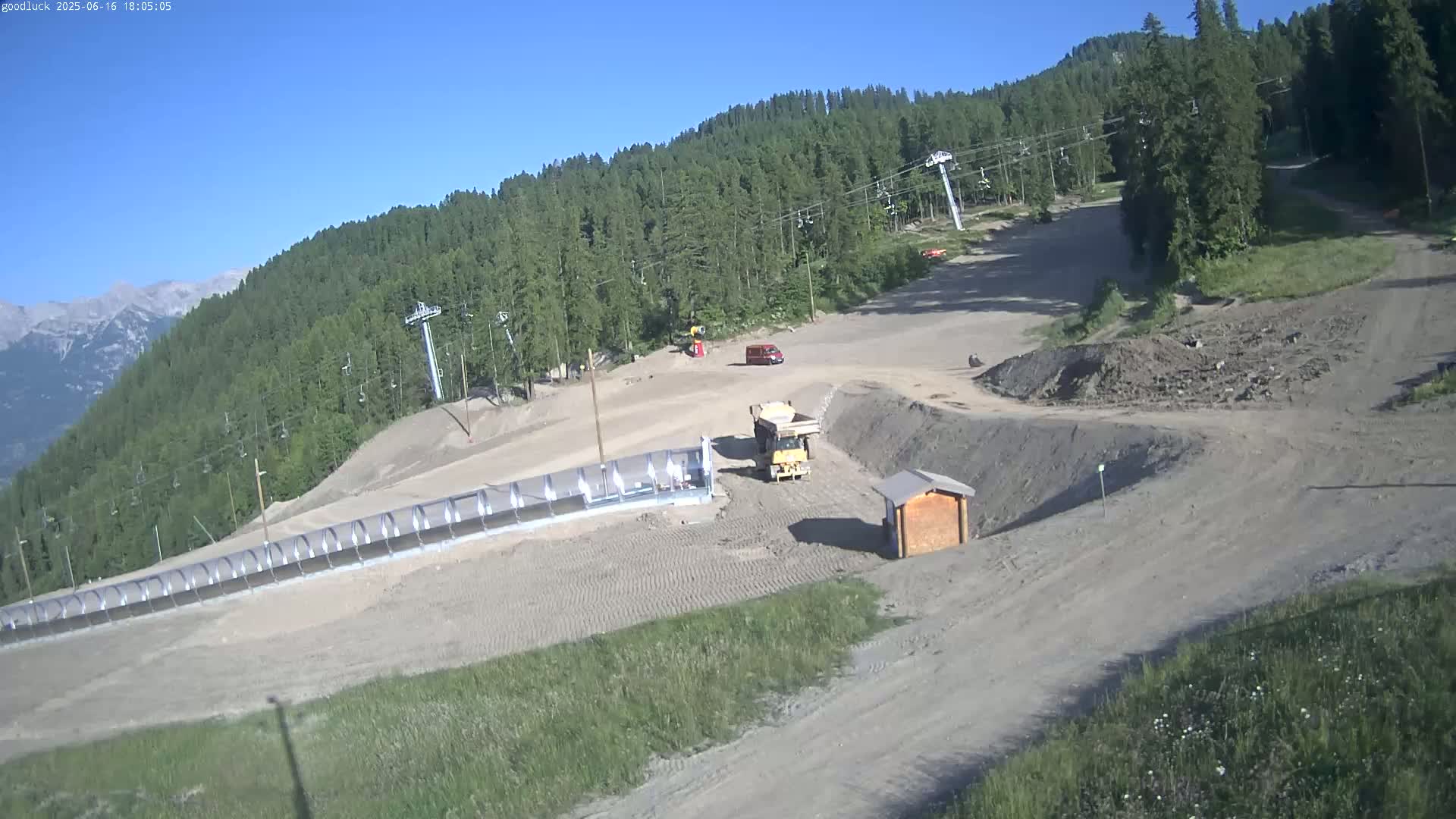 Under a clear sky, construction vehicles are visible on a mountainside ski slope with several chairlifts and a long protective barrier.