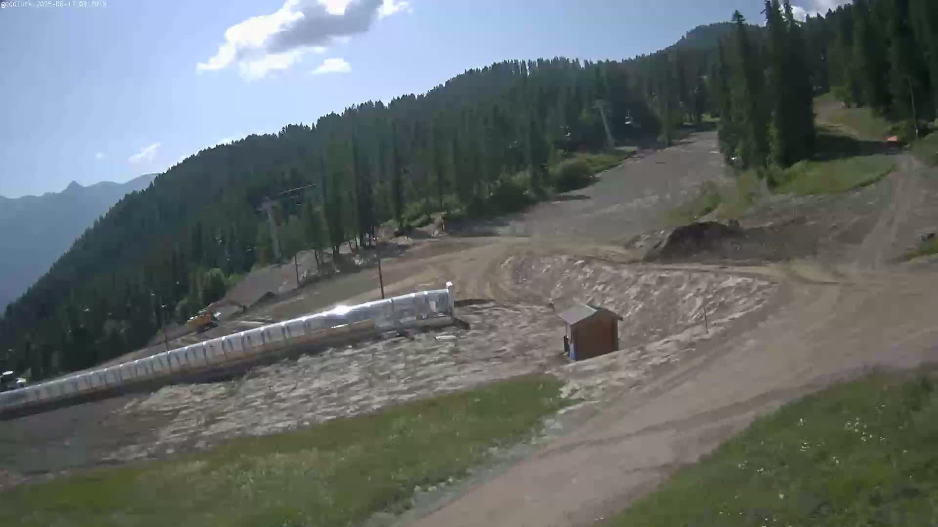 A mostly sunny view of a mountainside ski resort with a muddy, mostly cleared ski run and a long, covered ski lift in the foreground.