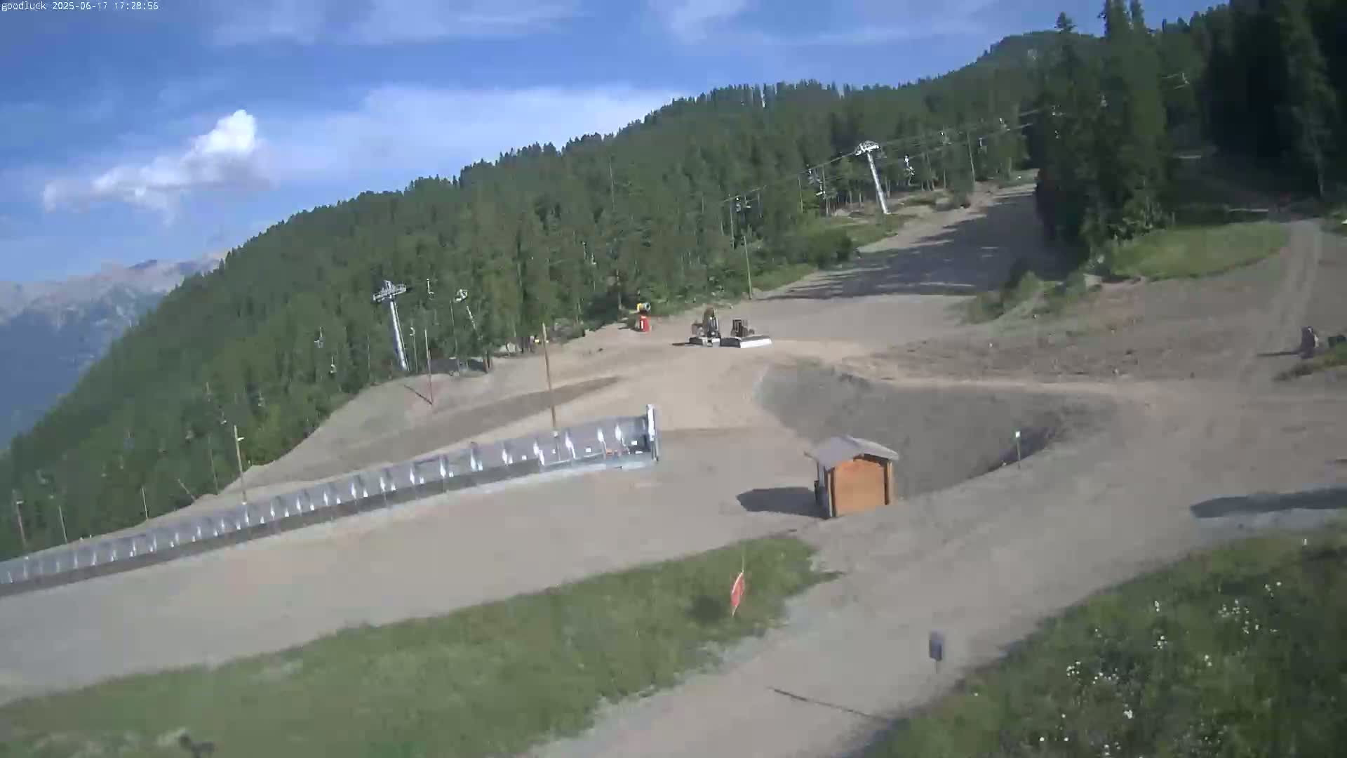 A mostly sunny day reveals a ski slope in the summer, with exposed dirt and sparse vegetation, featuring a snow-grooming machine and chairlift infrastructure.