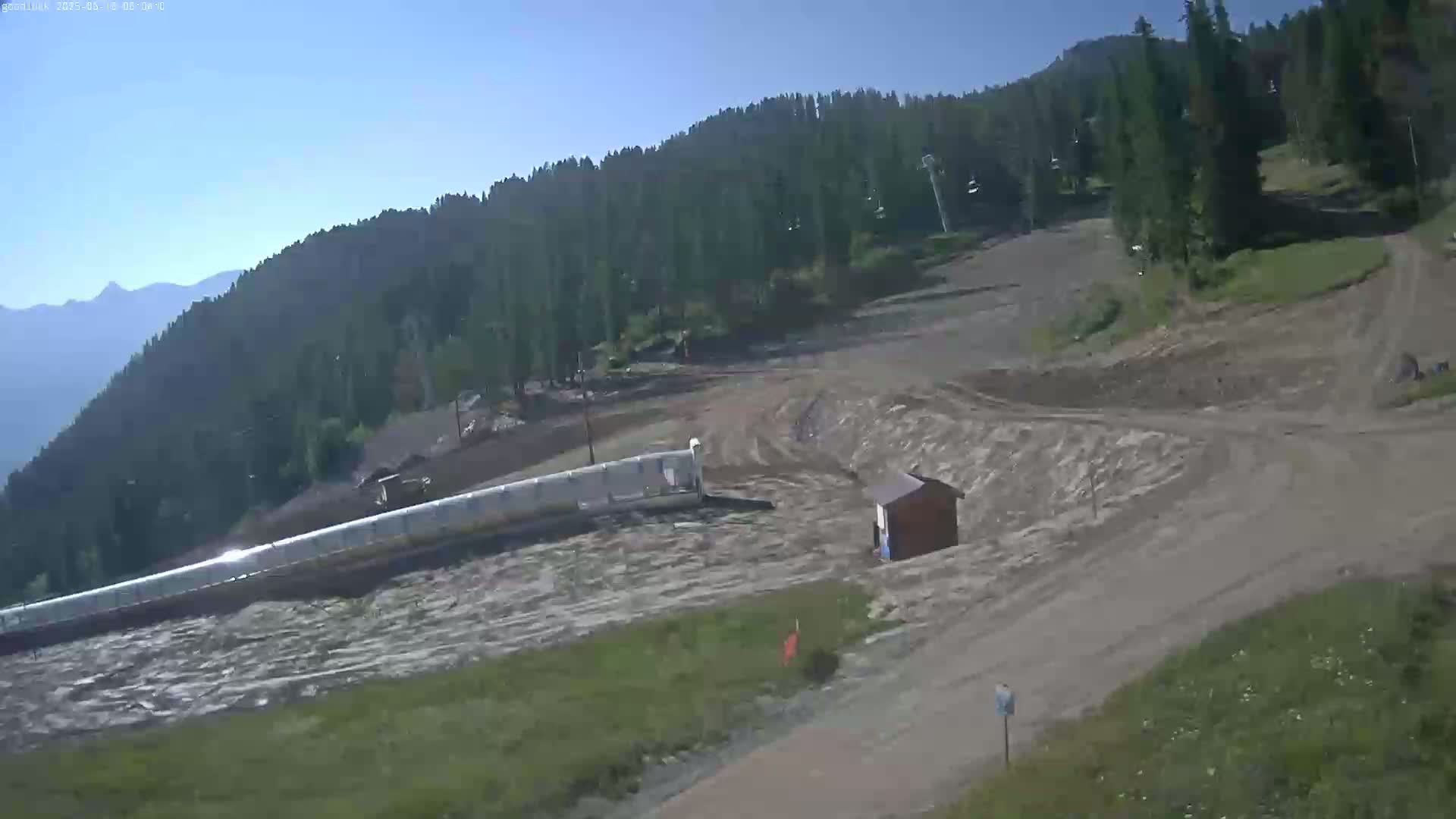 A mostly clear, sunny day reveals a mountainside ski area undergoing apparent construction or maintenance, with muddy ground and equipment visible.