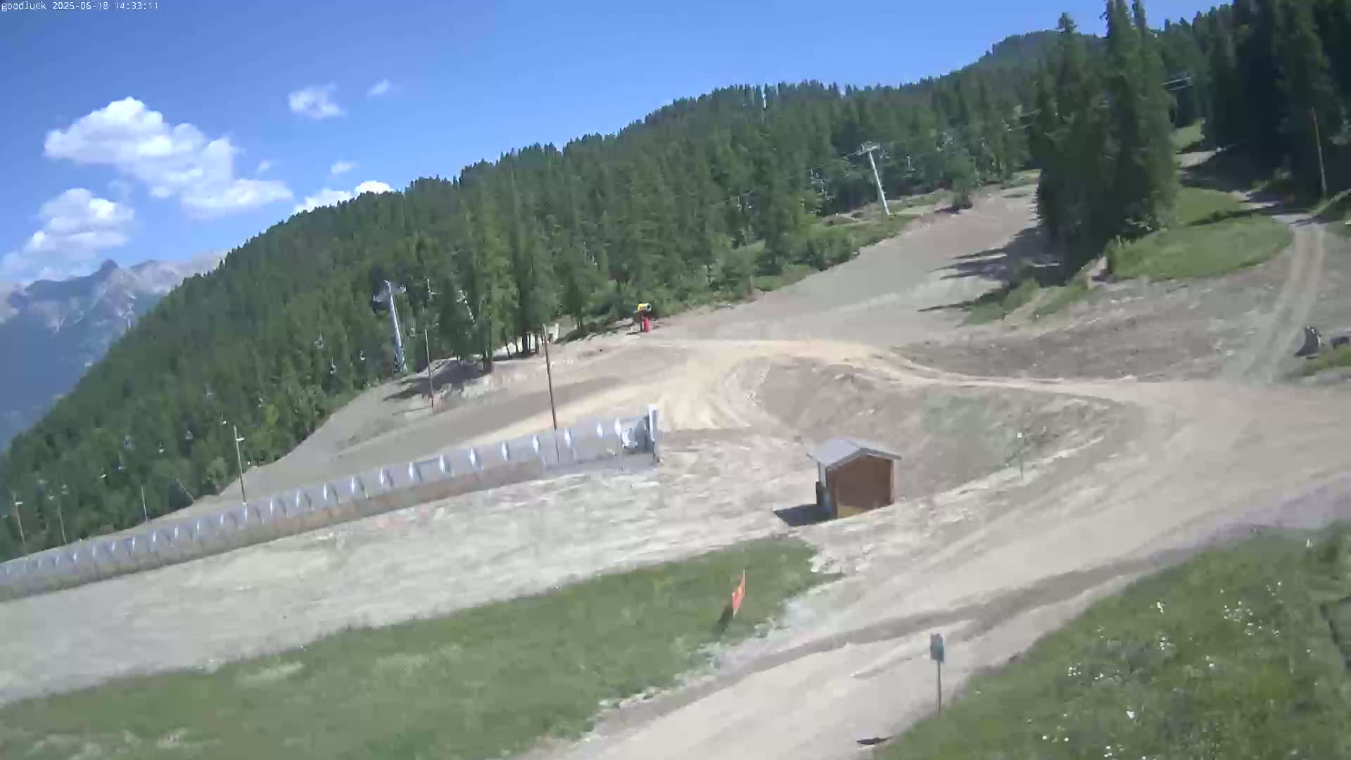 A mostly sunny day reveals a mountainside ski area with bare, dirt paths winding through sparse vegetation and a forested hillside.
