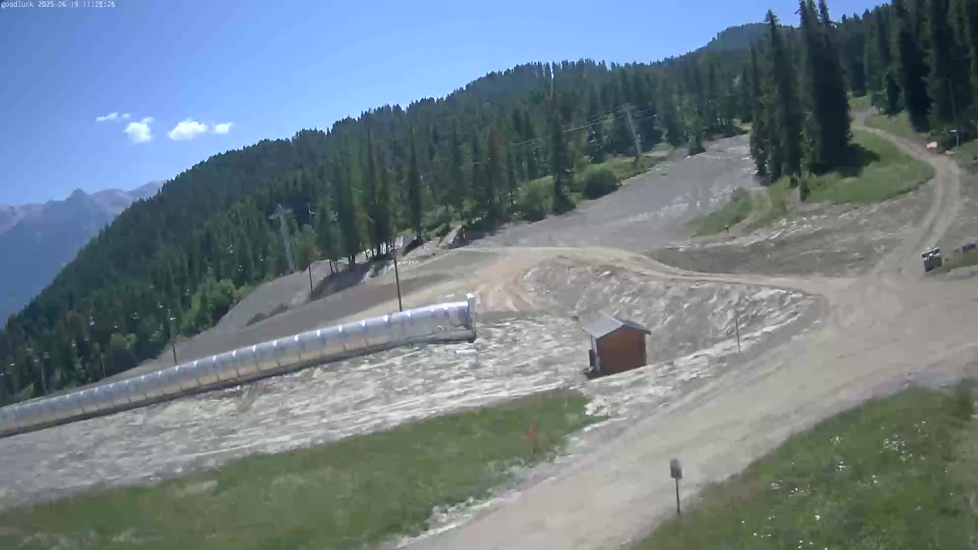 A high-angle view shows a mountainous area with a ski slope, a long, covered structure, and a small building under a clear, sunny sky.