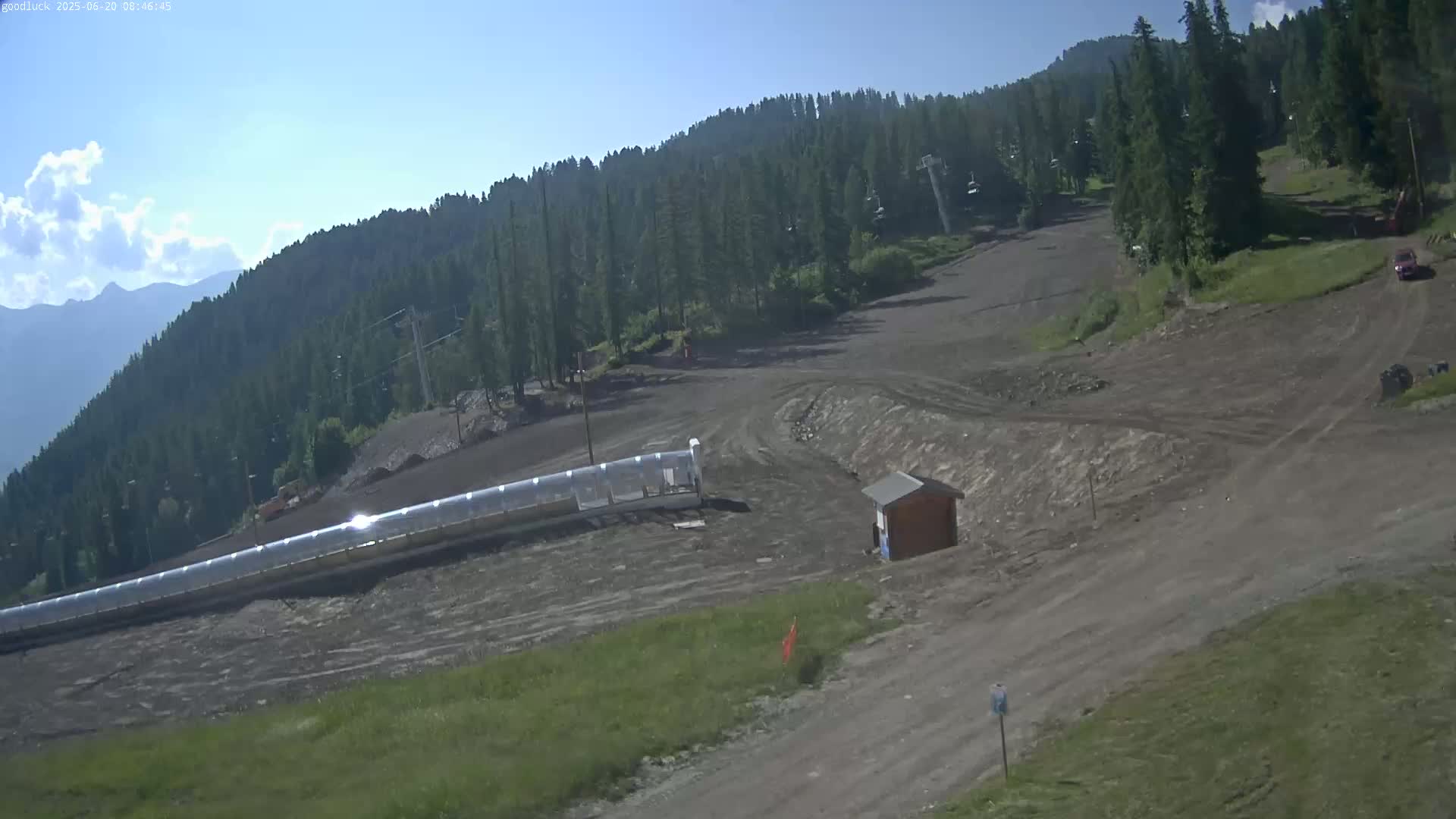 A mostly sunny day shows a large, mostly barren ski slope with a long, covered toboggan run, a small building, and a mountain range in the background.