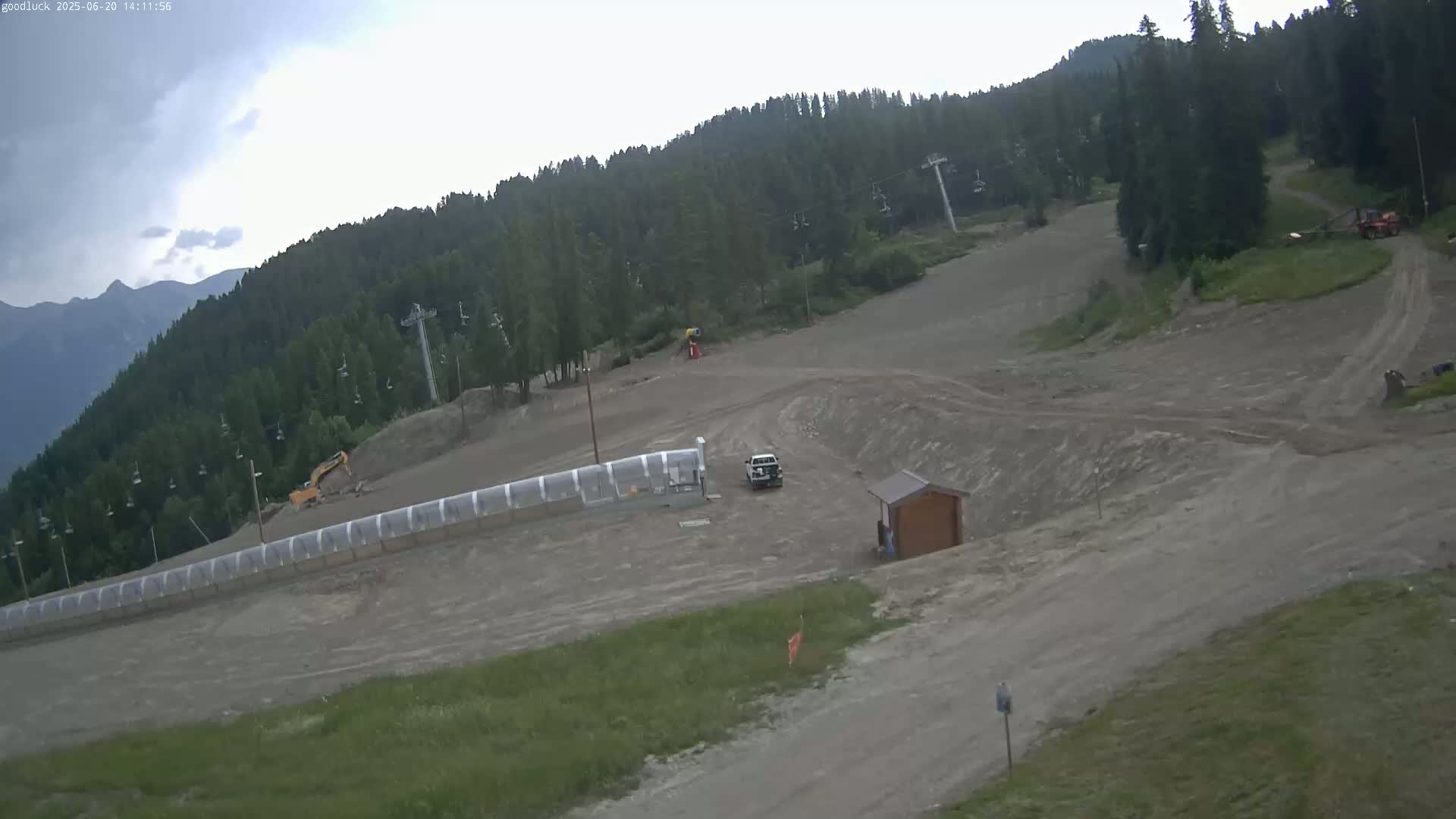A mostly cloudy day reveals a ski slope under construction, with various machinery and vehicles present, surrounded by a dense forest and mountains in the distance.