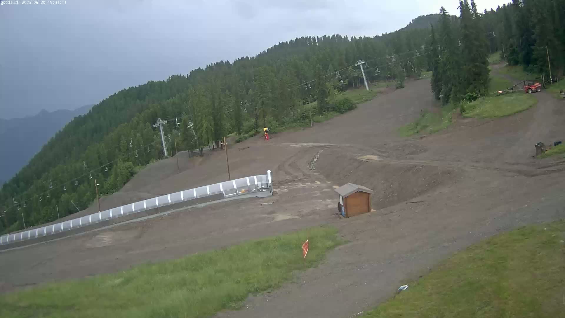 A mostly overcast day shows a ski slope in the summer, with exposed dirt and a few pieces of equipment visible, surrounded by a green forest.