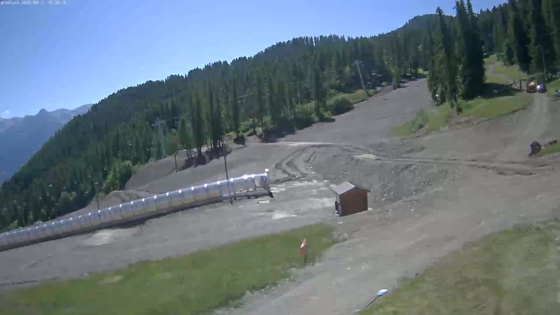 A mostly sunny, dry mountaintop ski slope shows a long, covered conveyor belt, a small building, and tire tracks in the dirt.