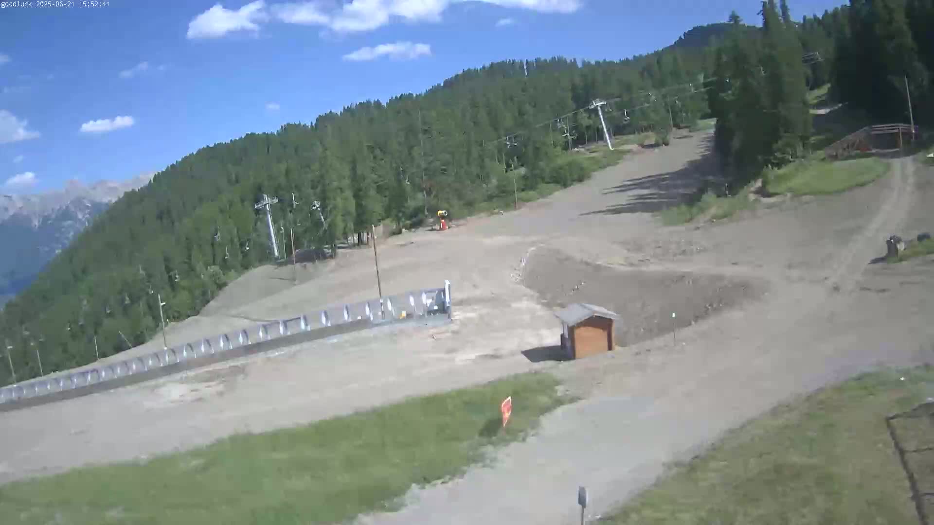 A mostly sunny day reveals a ski slope under construction, surrounded by a lush green forest and distant mountains.