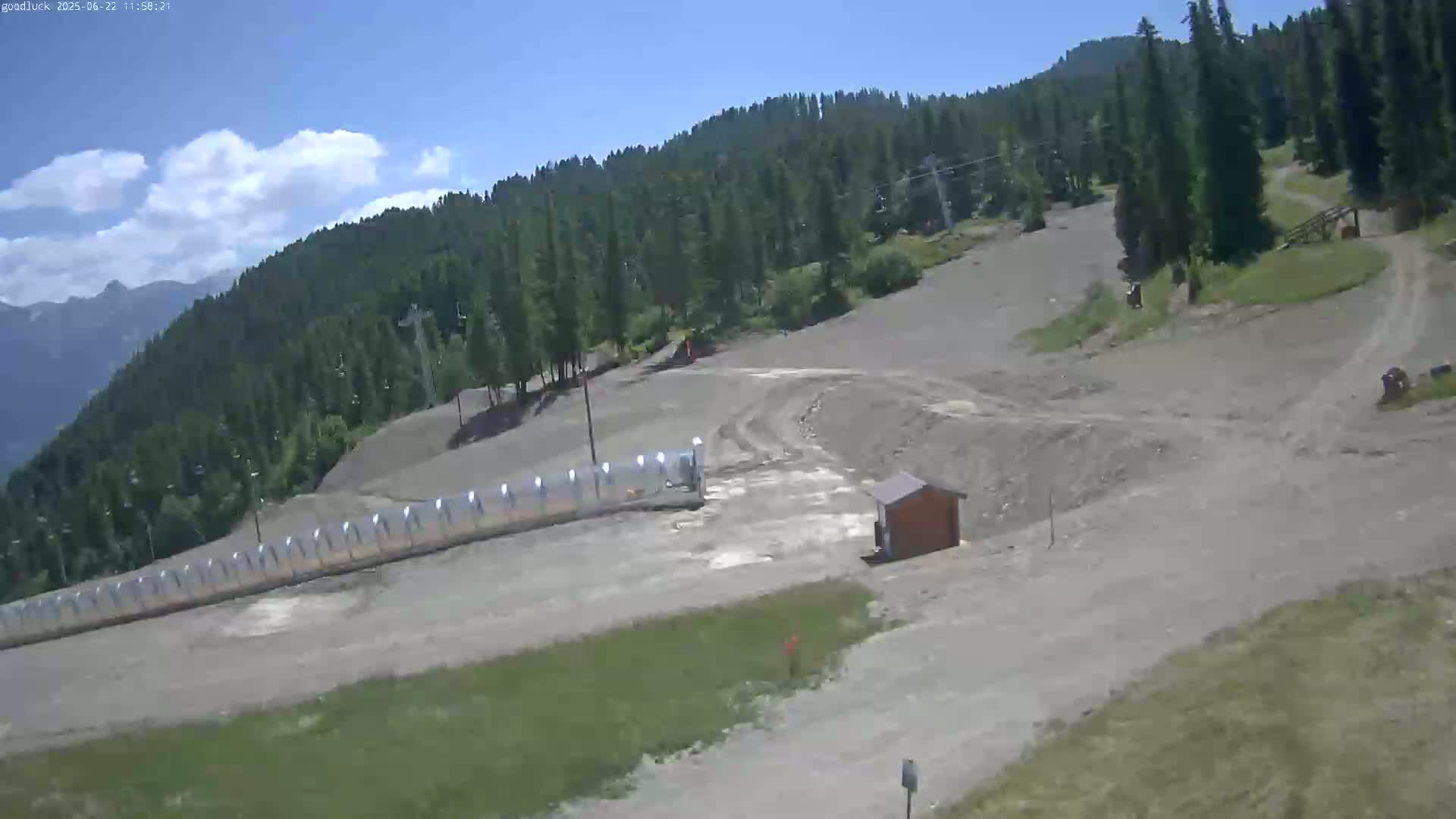 A mostly clear, sunny day reveals a mountainside ski slope with a small building and a long, covered structure, surrounded by evergreen trees.