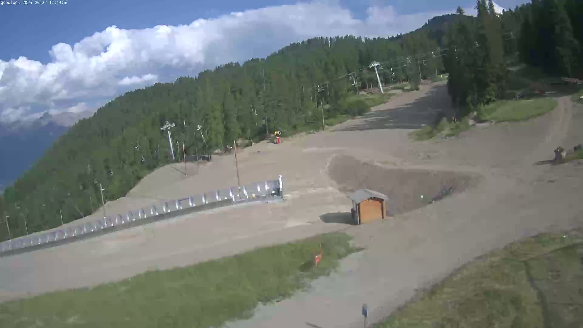 A mostly sunny, daytime view of a ski slope in the summer, showing a mostly barren slope with some sparse vegetation, a small building, and ski lifts in the background with a mountain range visible in the distance.
