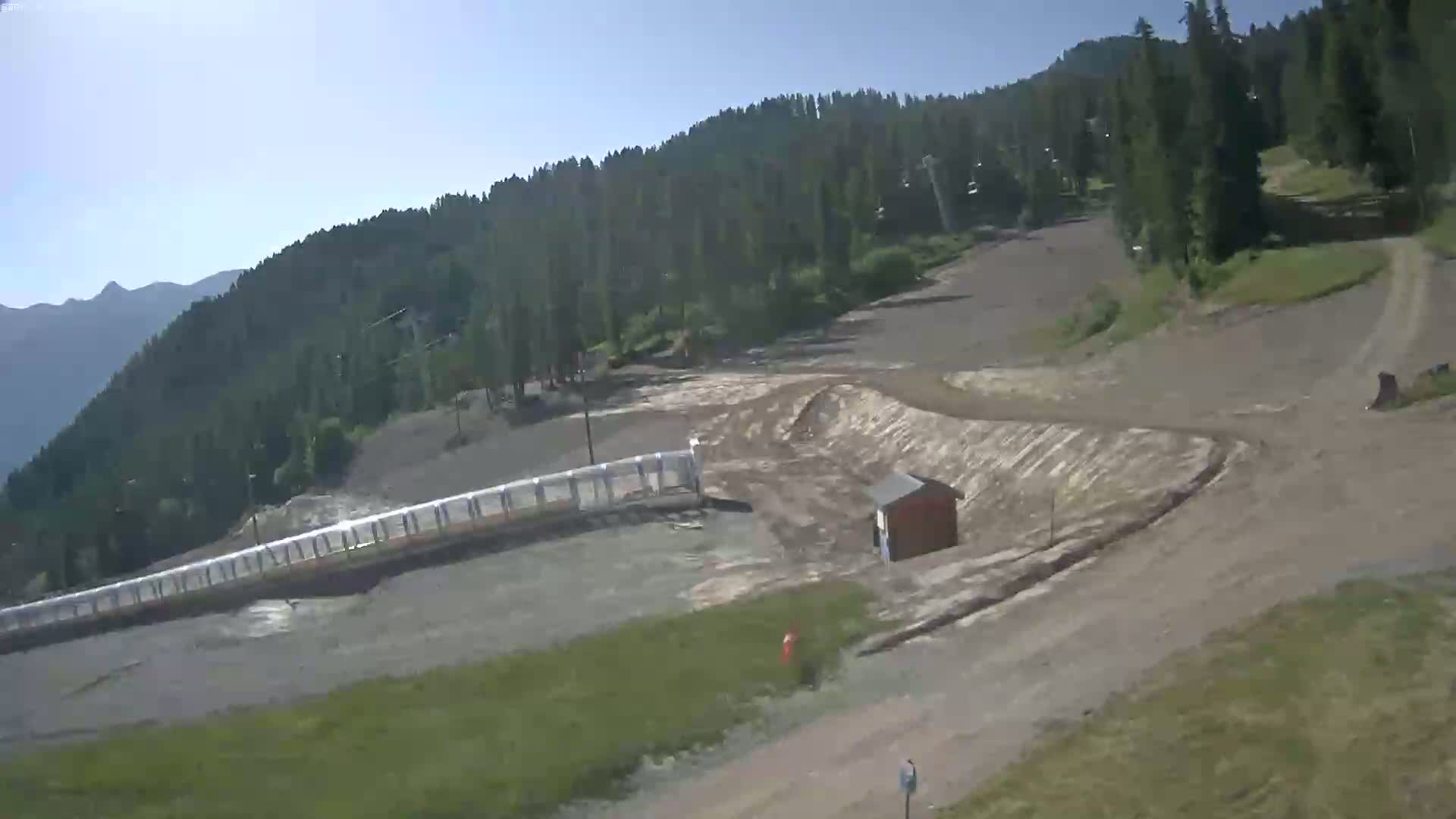 A mostly sunny view of a mountainous ski resort during the summer shows a dirt path winding through a grassy area, next to a long, low protective barrier and a small building.