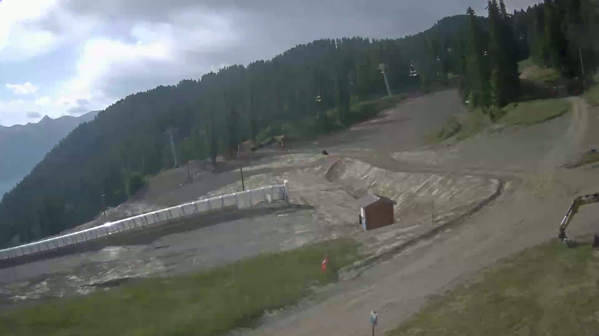 A mostly cloudy day reveals a mountainside ski resort under construction, showing dirt paths, a small building, and earth-moving equipment.