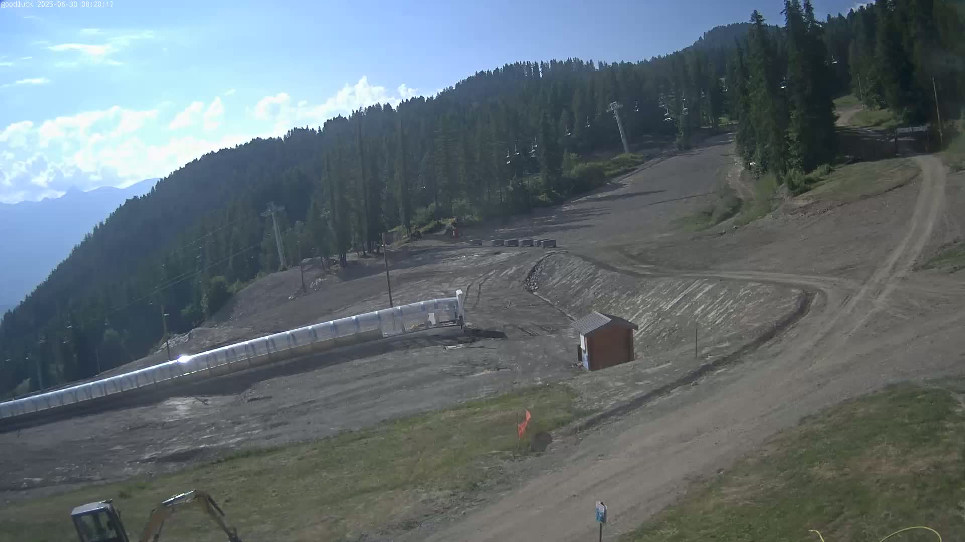 A mostly sunny day reveals a ski slope in the summer, showing bare ground, a long enclosed lift, and some small structures.