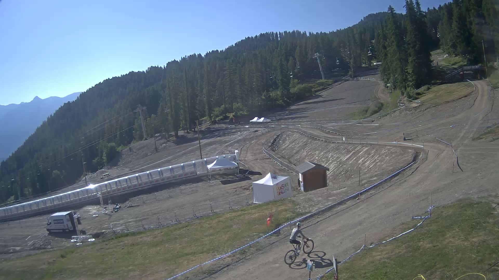A mountain biker rides a trail on a sunny day, amidst a dirt course surrounded by a forested hillside.