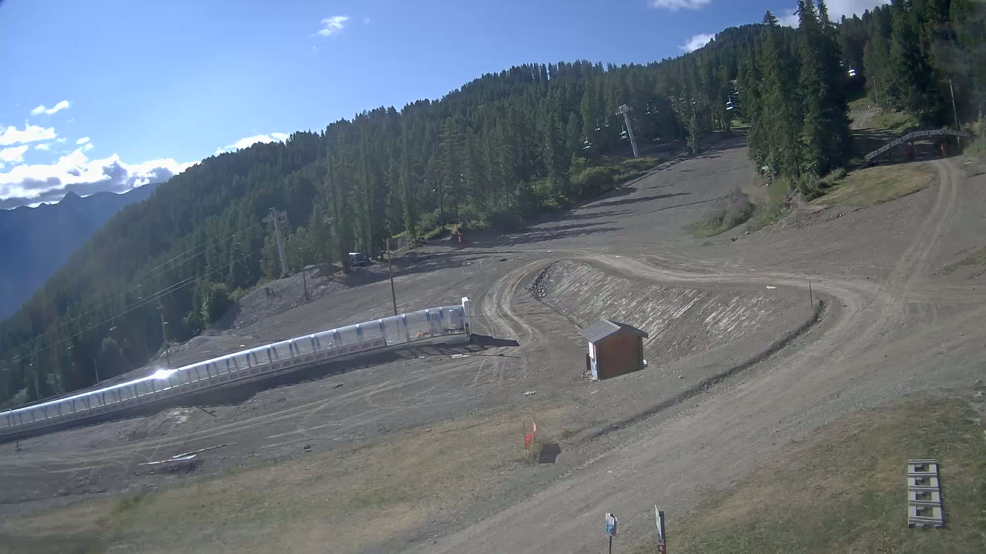 A mostly sunny day reveals a mountainside ski area with dirt paths, a long covered conveyor lift, and a small building, all surrounded by evergreen trees.