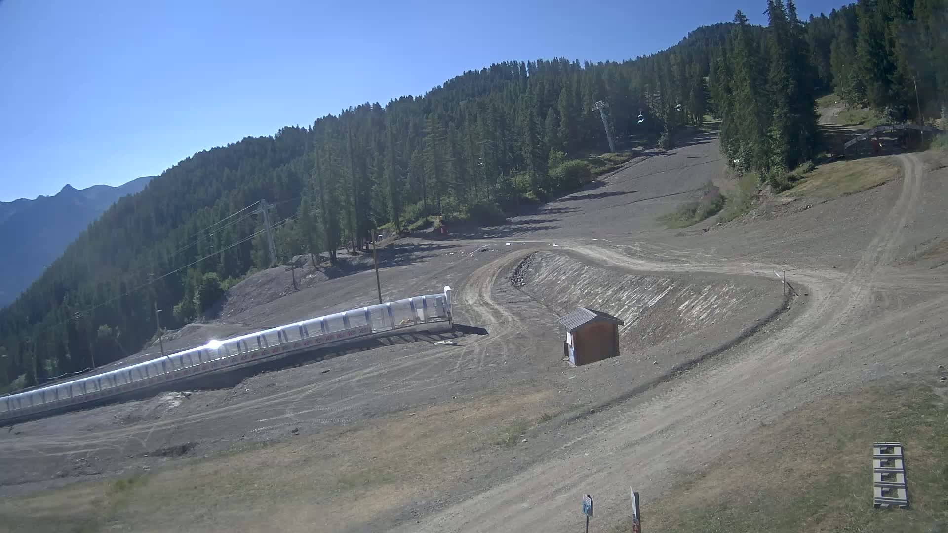 A mostly barren ski slope with a long, transparent protective barrier at the bottom, under clear, sunny skies, is surrounded by a dense evergreen forest and distant mountains.