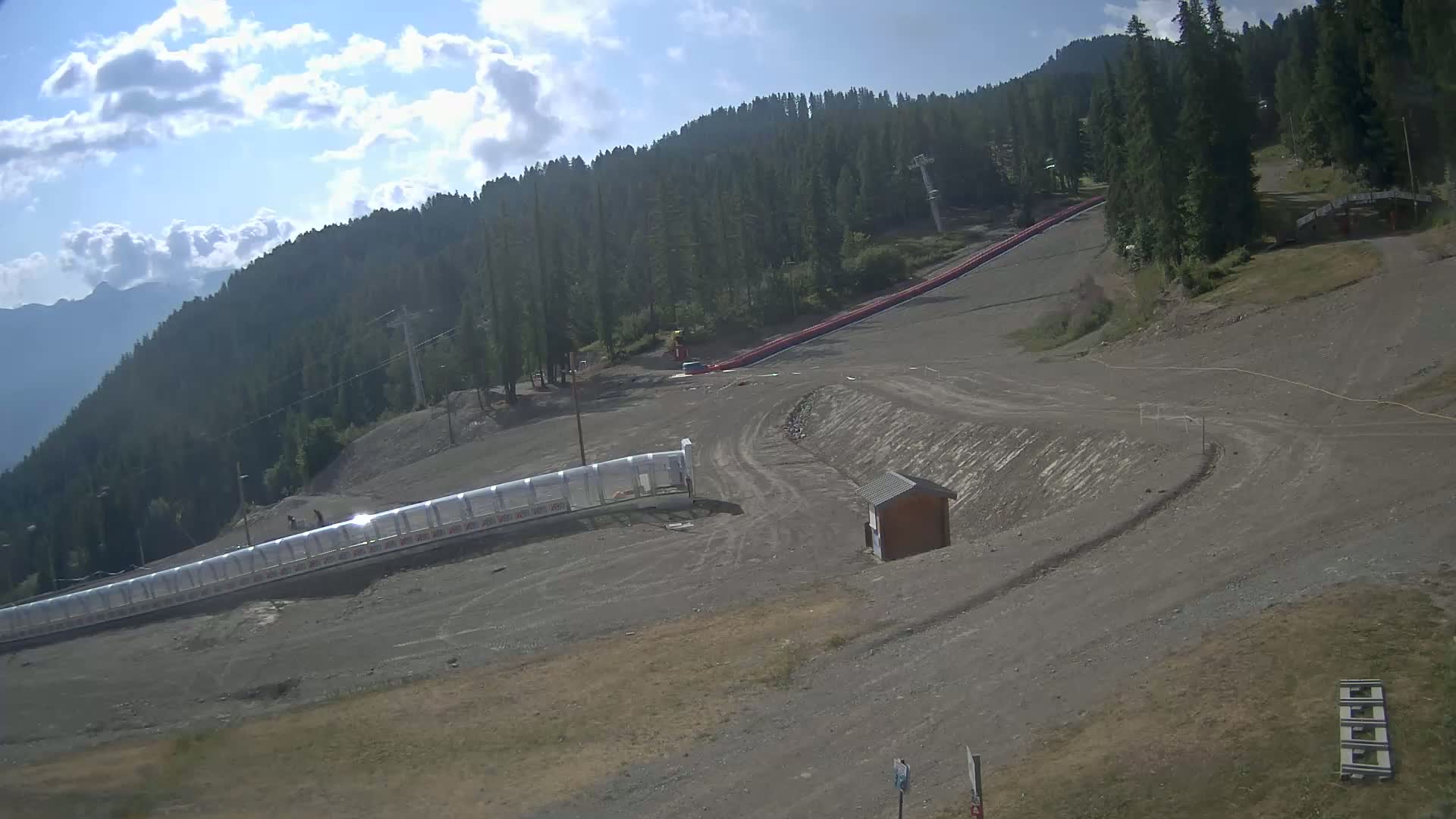 A mostly sunny mountainside with a long, red tubing hill, a clear plastic enclosure, and a small wooden building.