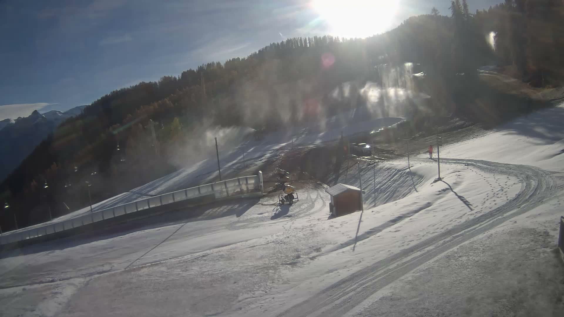On a bright, sunny day, snow cannons are actively producing snow on a partially covered ski slope, with a person walking on a snowy path, backed by wooded mountains and distant peaks under a clear blue sky.