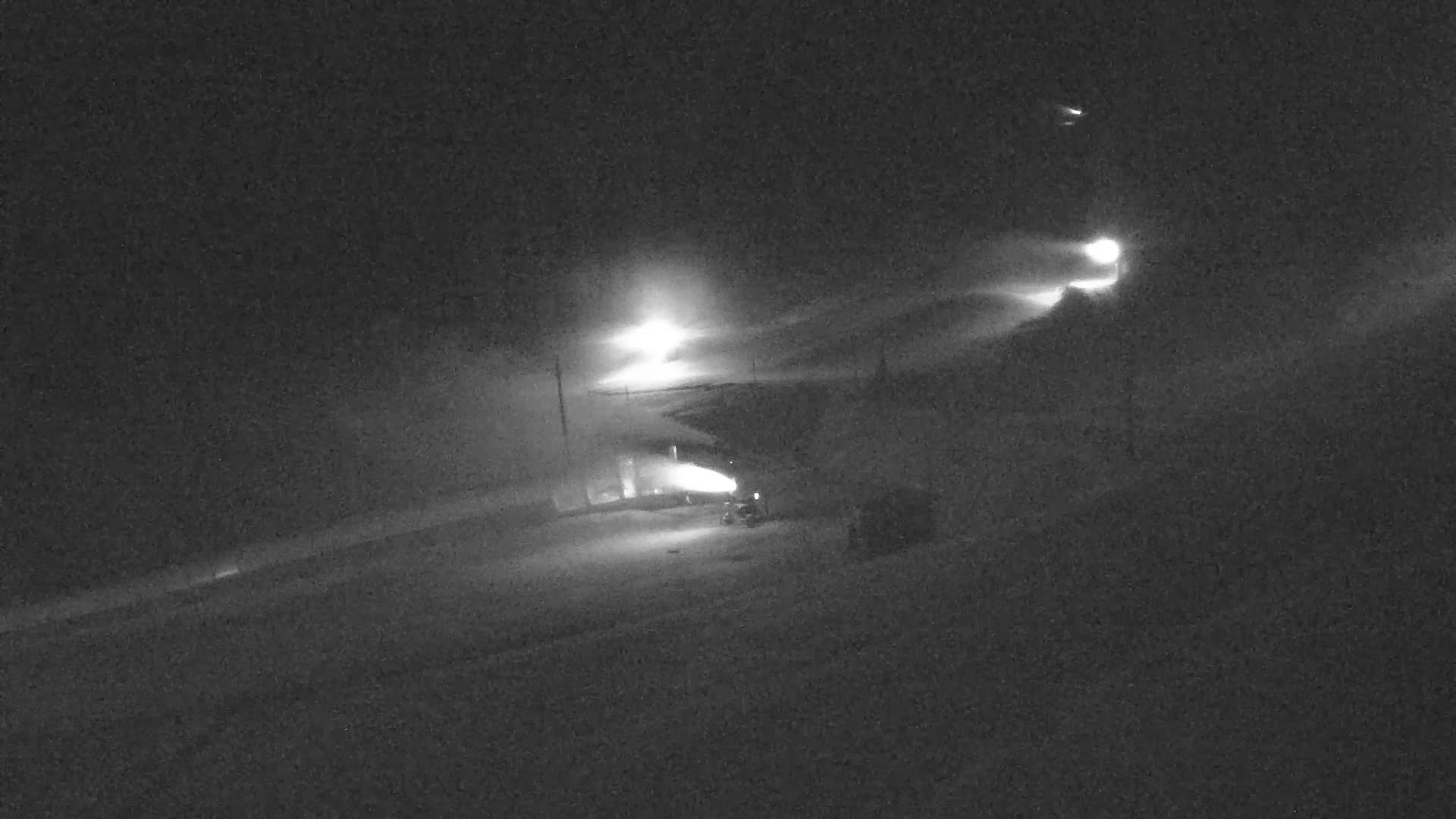 On a bright, sunny day, snow cannons are actively producing snow on a partially covered ski slope, with a person walking on a snowy path, backed by wooded mountains and distant peaks under a clear blue sky.