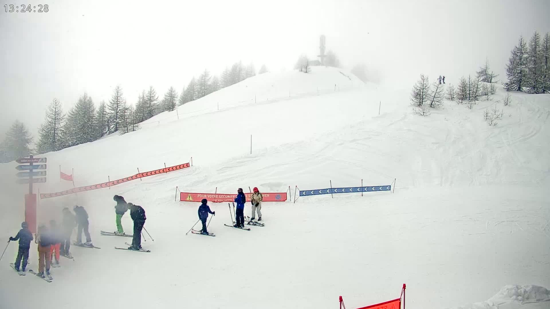 A snowy ski slope on a foggy day has several skiers and snowboarders at the bottom, with a few more visible higher up the hill.