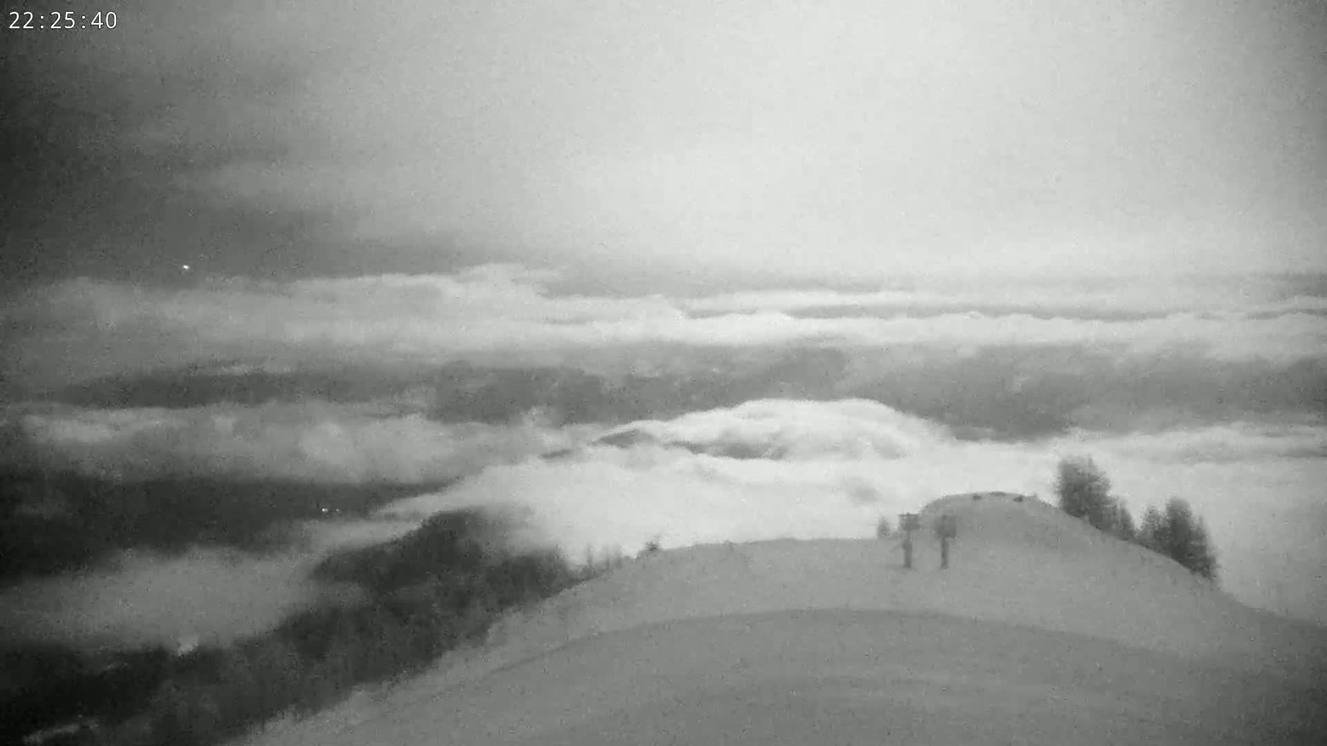 A high-angle, nighttime view shows two figures standing on a snow-covered hilltop overlooking a vast expanse of clouds under a gray sky.