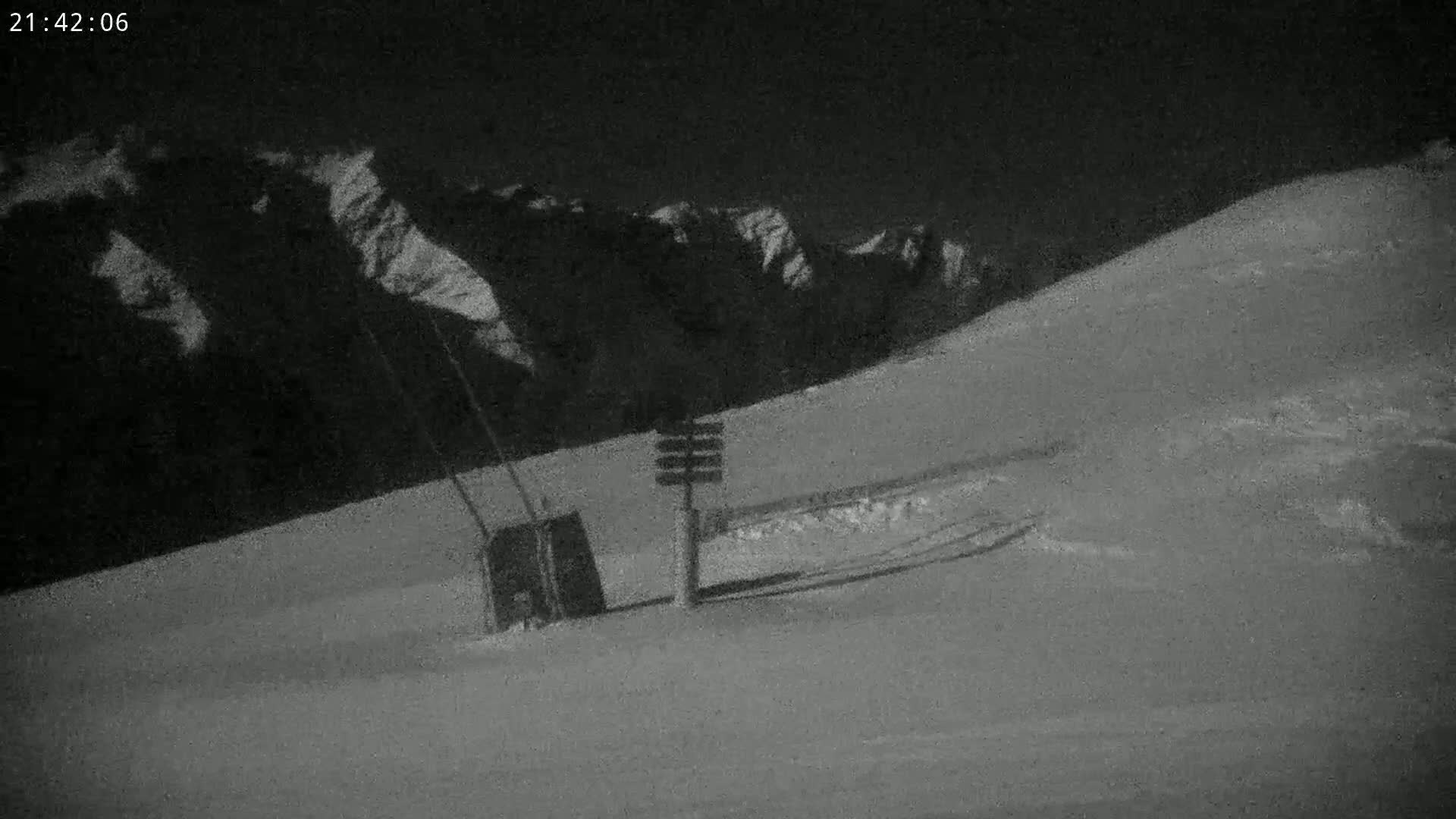 A nighttime, snow-covered slope with a ski lift and signage is visible under a dark sky, mountains in the background.