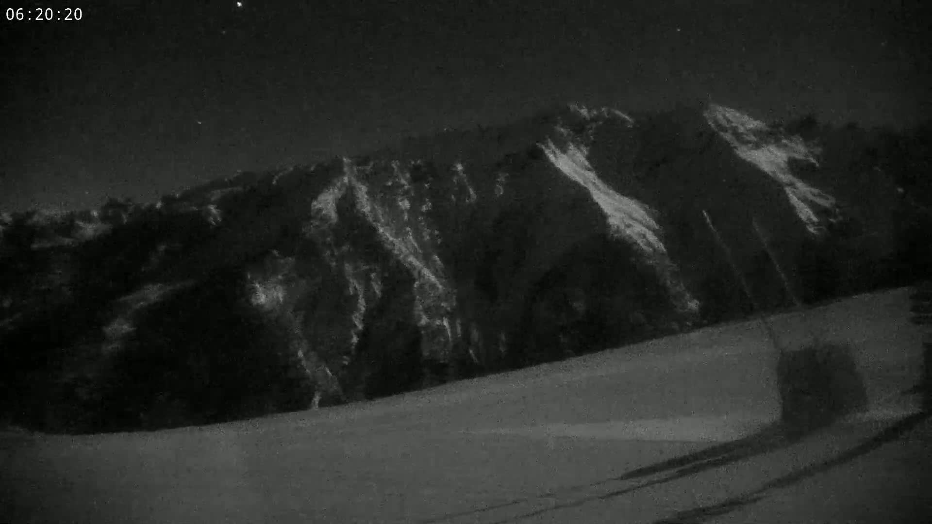 A nighttime, low-light image shows a snow-covered landscape with a dark mountain in the background under a clear, starry sky.