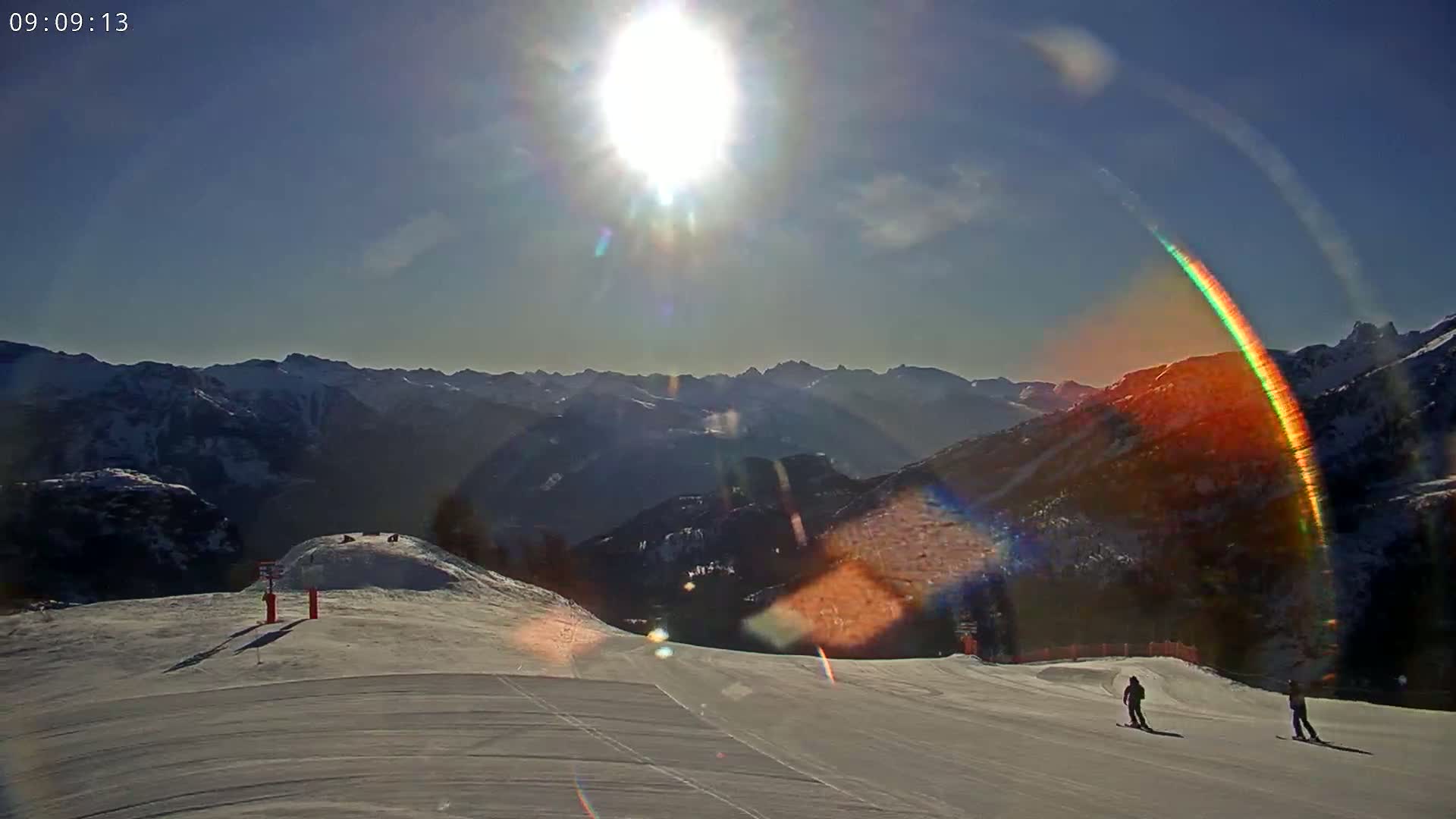 A sunny day on a snow-covered mountain slope shows two skiers on a wide run, with a mountain range in the background.