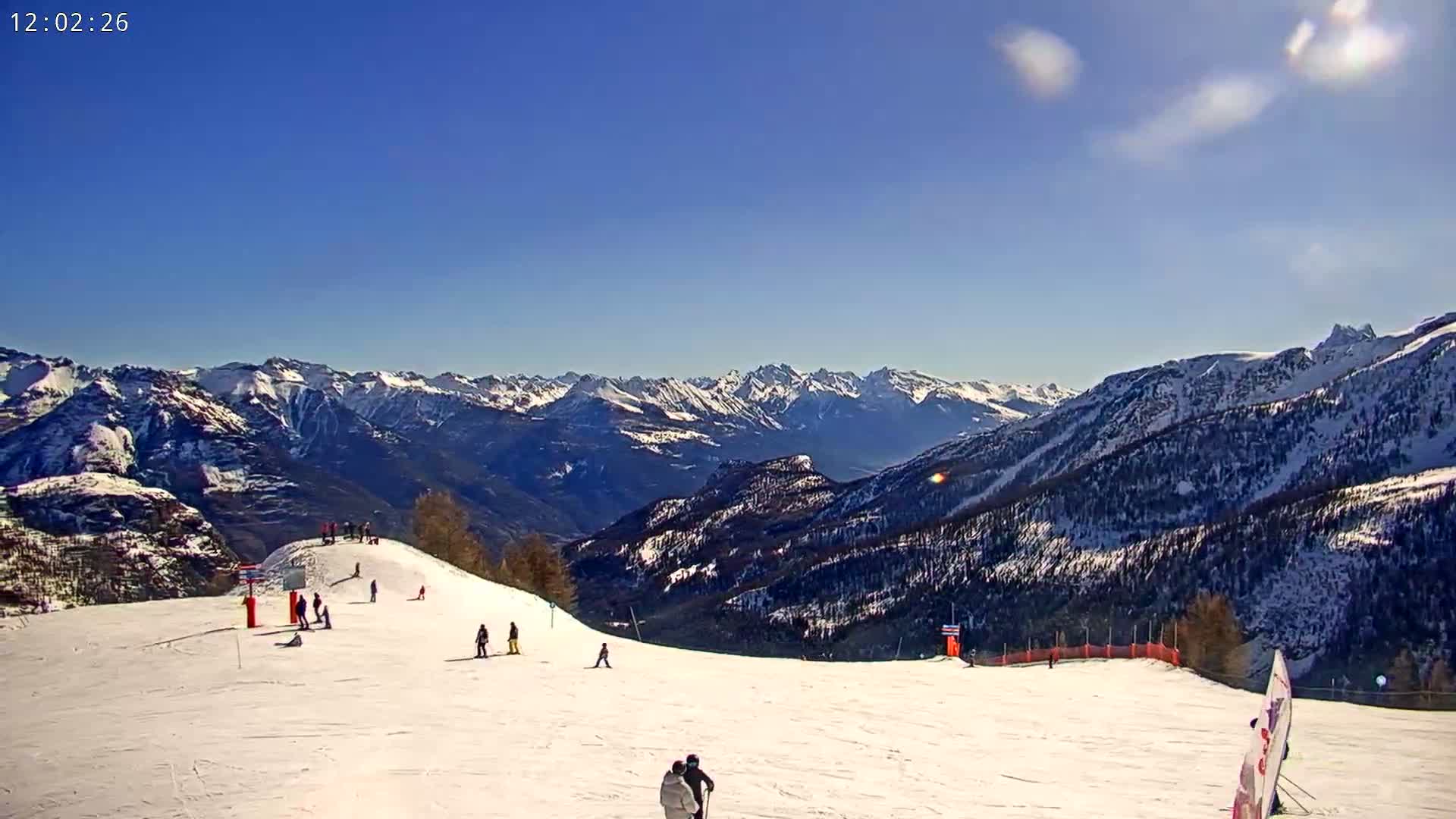 A sunny day on a snow-covered ski slope overlooking a vast mountain range.