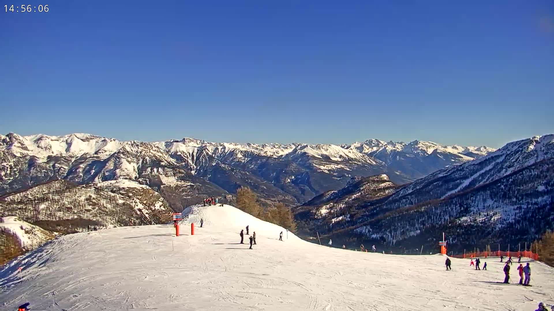 A snow-covered ski slope with skiers and snowboarders, situated in a mountainous region under a clear blue sky.